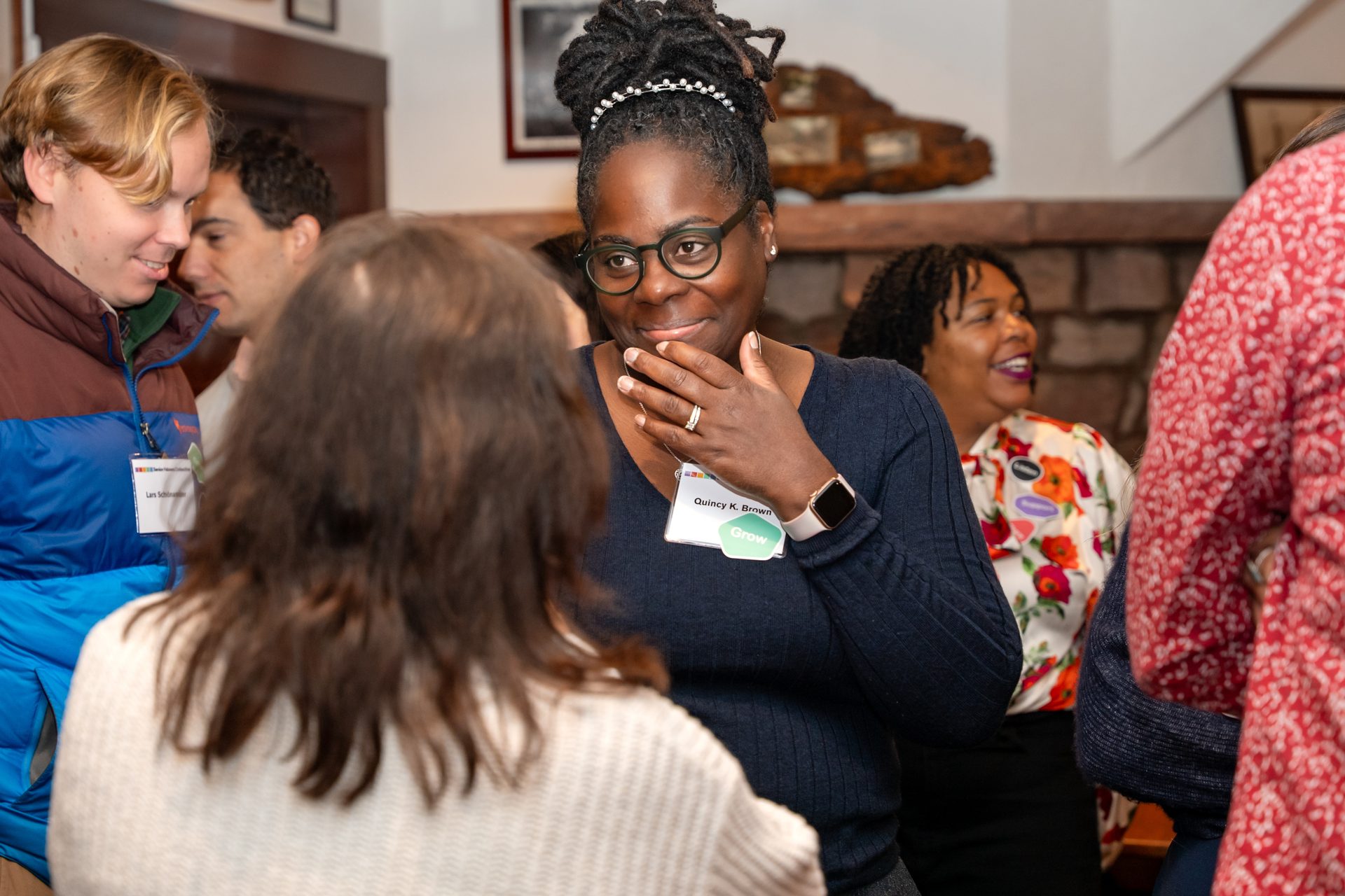 Quincy K. Brown, a Black woman with glasses, smiles and holds her chin at a networking event.