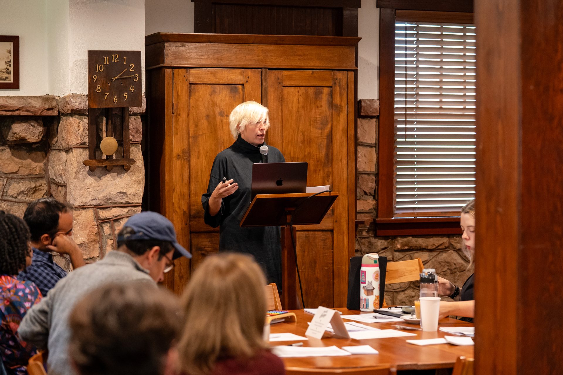 Blonde woman speaking at a podium with a laptop to an audience in a rustic room.