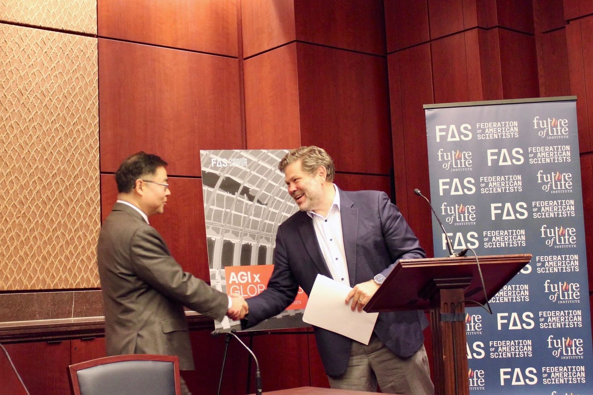Two men shake hands at a formal event, with "FAS" and "Future of Life Institute" banners behind them.