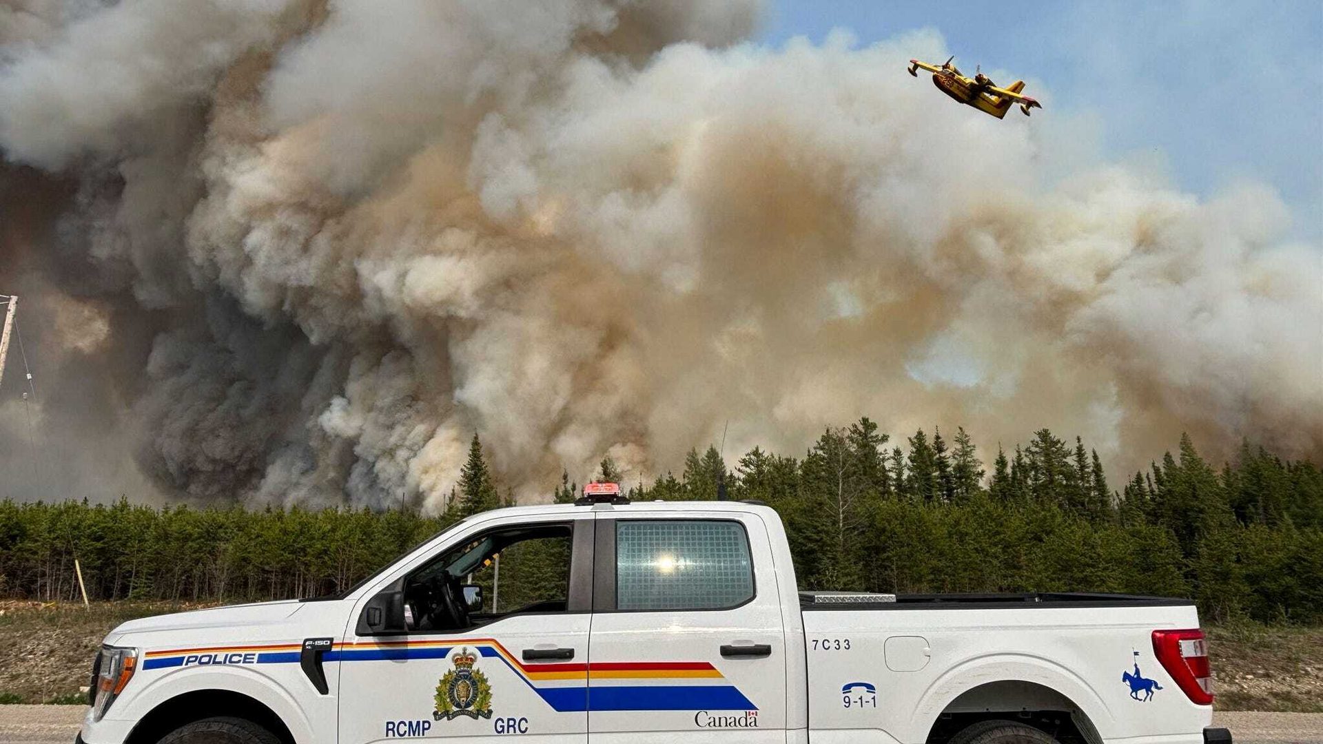 RCMP truck and a yellow water bomber aircraft in front of a large forest fire with thick smoke.