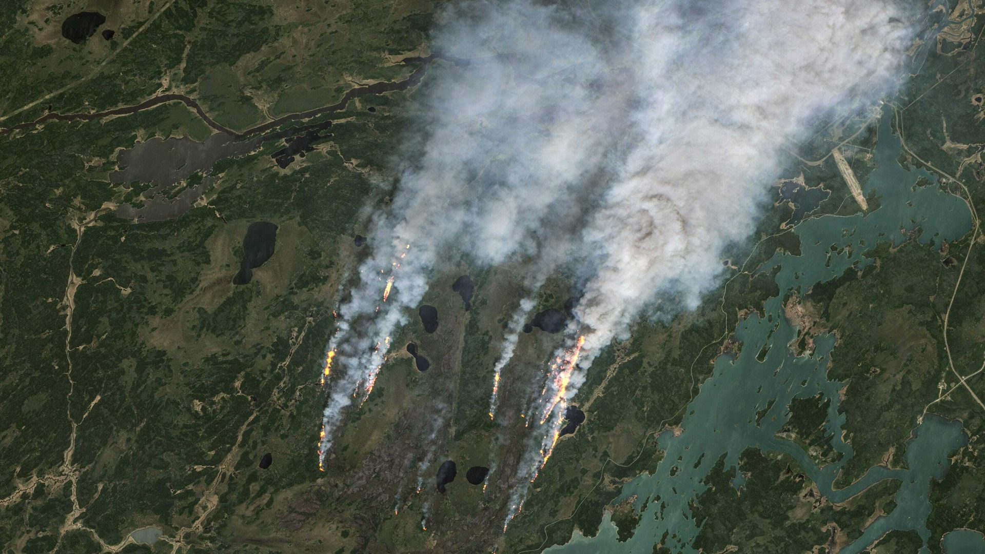 Aerial view of multiple wildfires with visible flames and dense smoke plumes over a forested landscape with lakes.