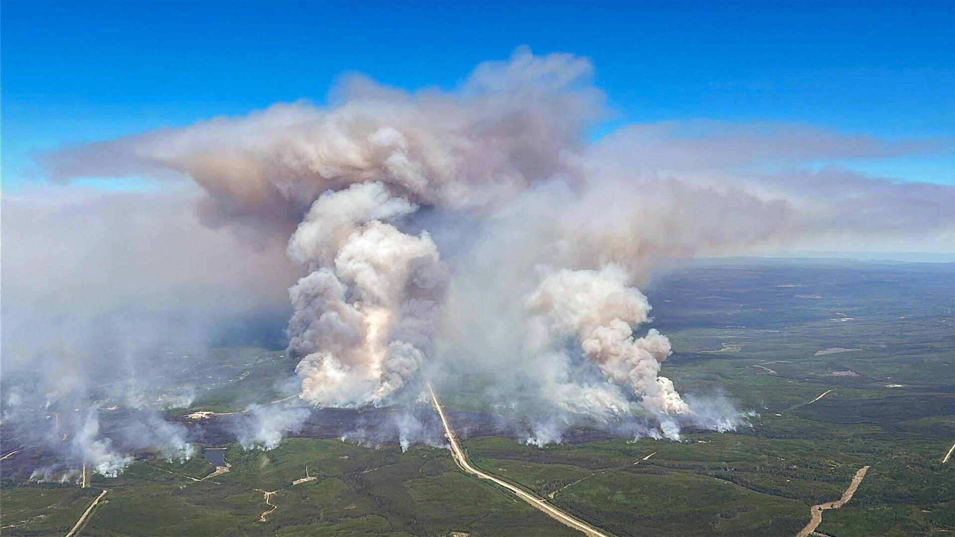 Huge wildfire: towering smoke plumes over a green forest with a road.