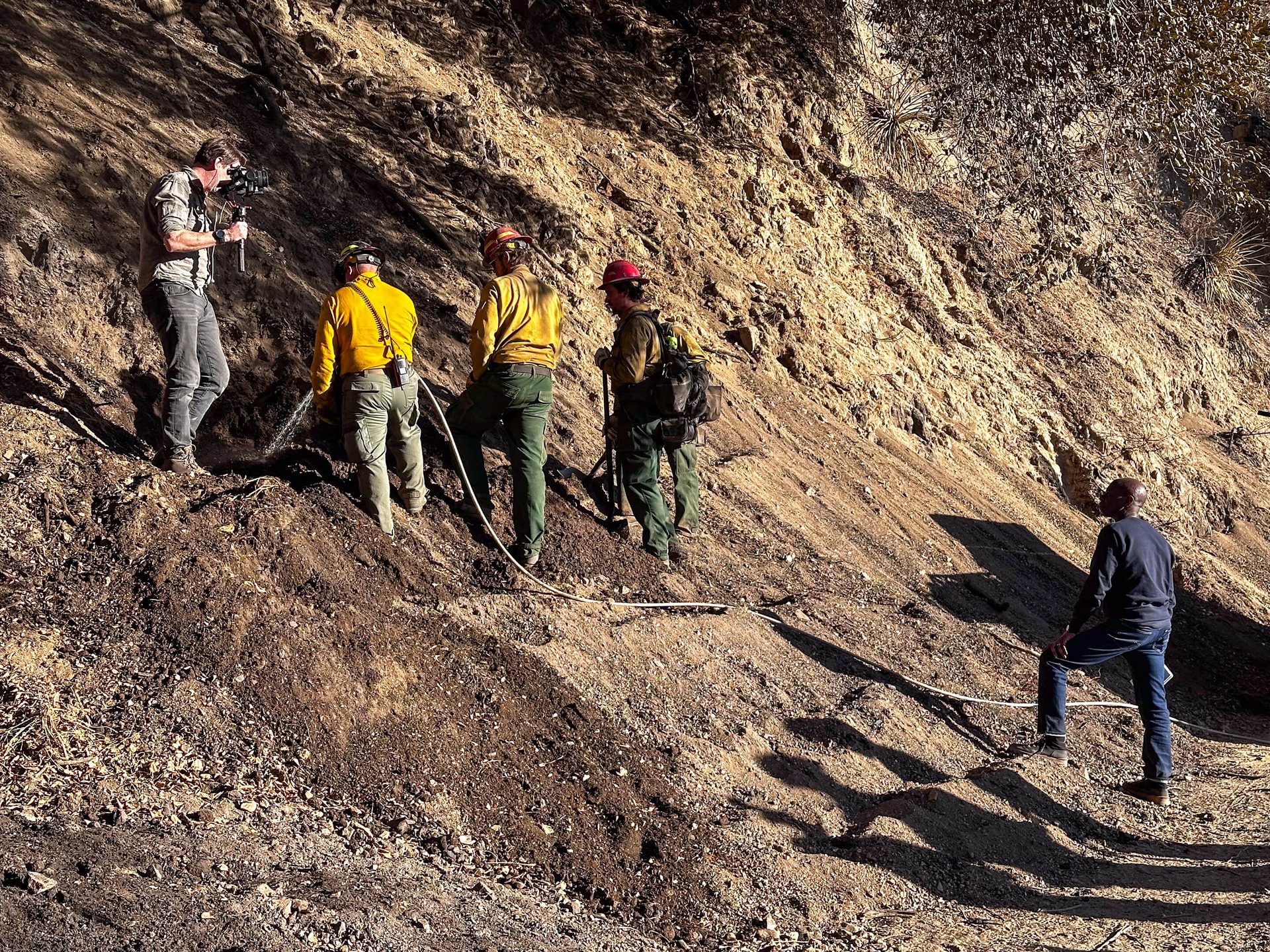 A filmmaker records firefighters spraying water on a steep dirt slope, while another man watches.
