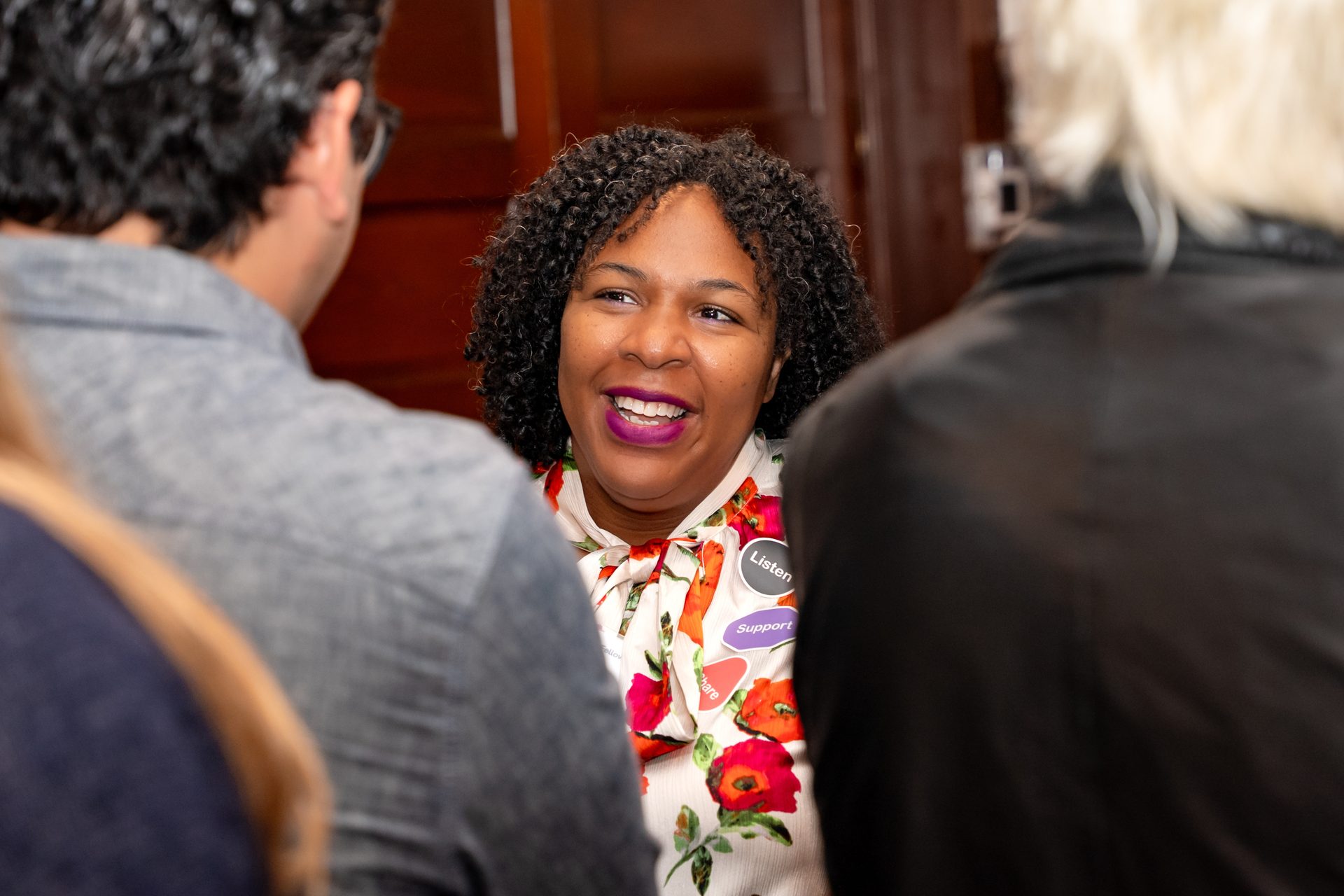 Smiling Black woman with curly hair, purple lipstick, "Listen" & "Support" badges, talking to others.