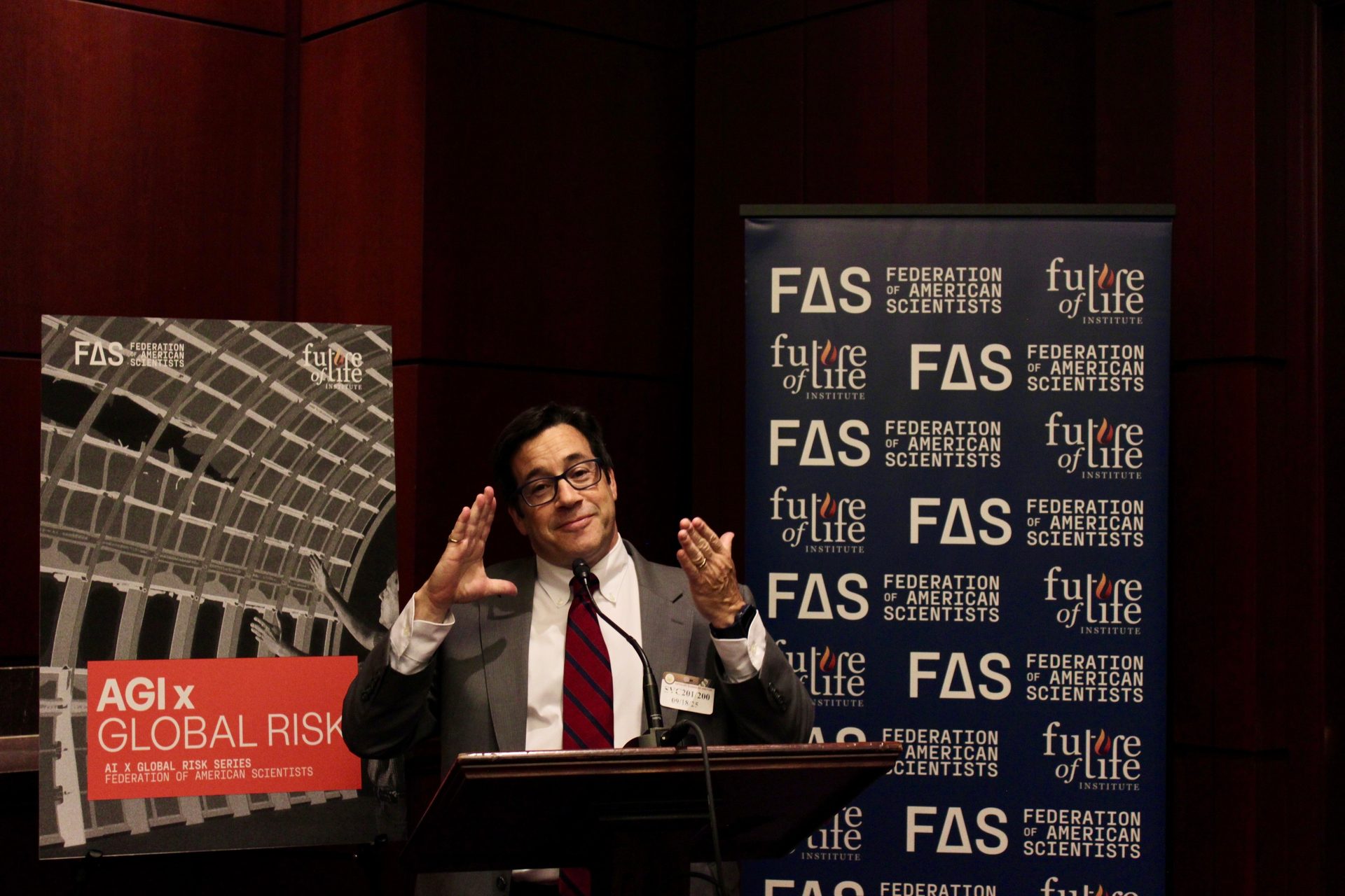 Man speaking at a podium, gesturing, with "FAS", "Future of Life", and "AGI x GLOBAL RISK" banners.