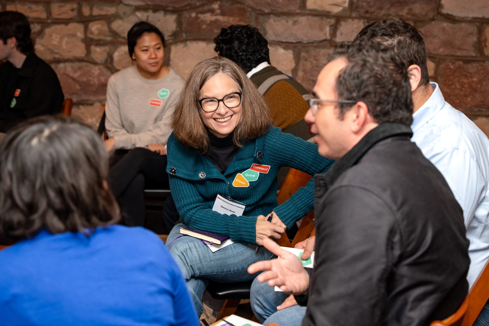 A woman in a teal sweater with "Connect, Grow, Create" badges smiles while talking in a group.
