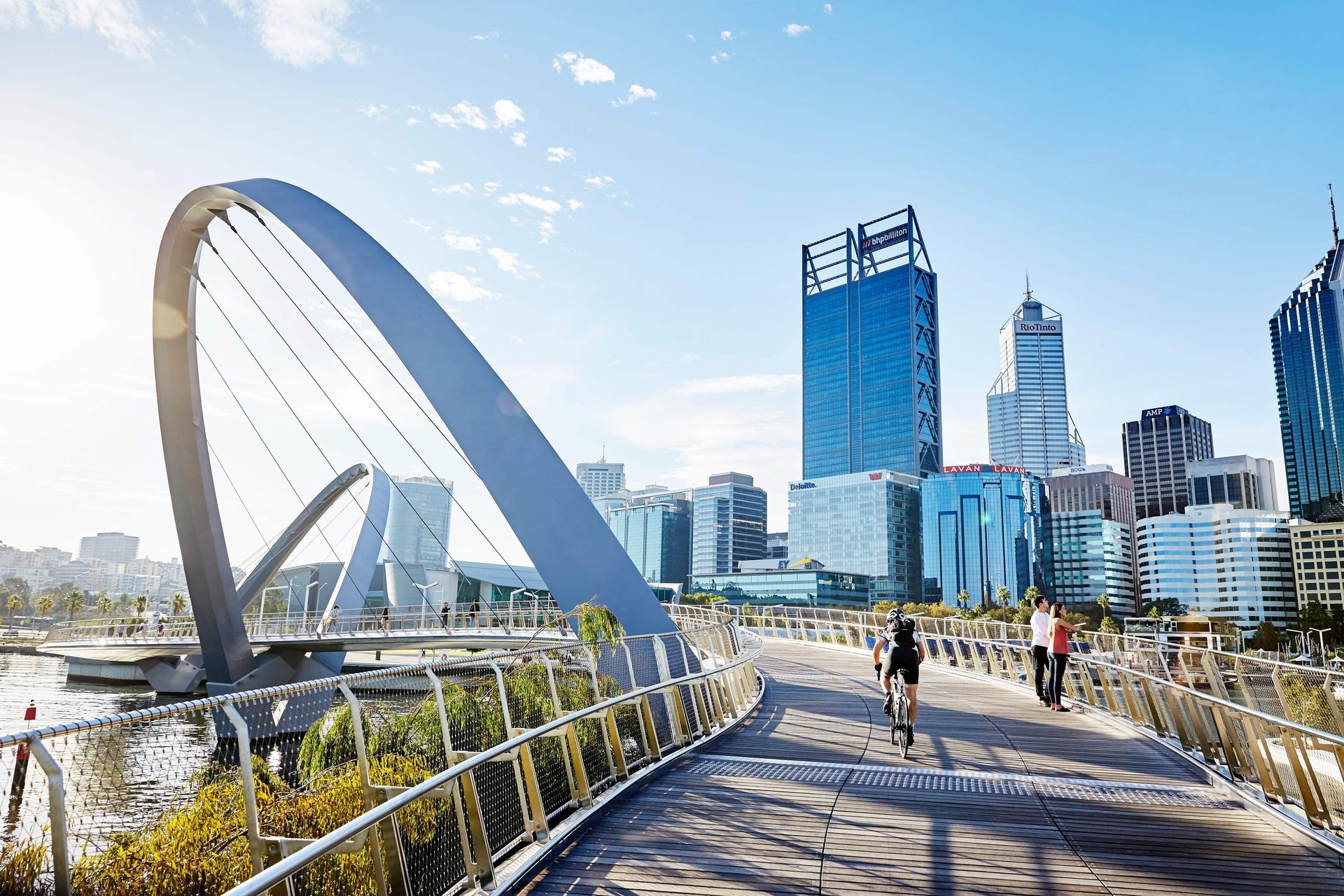 A modern pedestrian bridge, the Elizabeth Quay Bridge, curves towards Perth's city skyline under a clear sky.