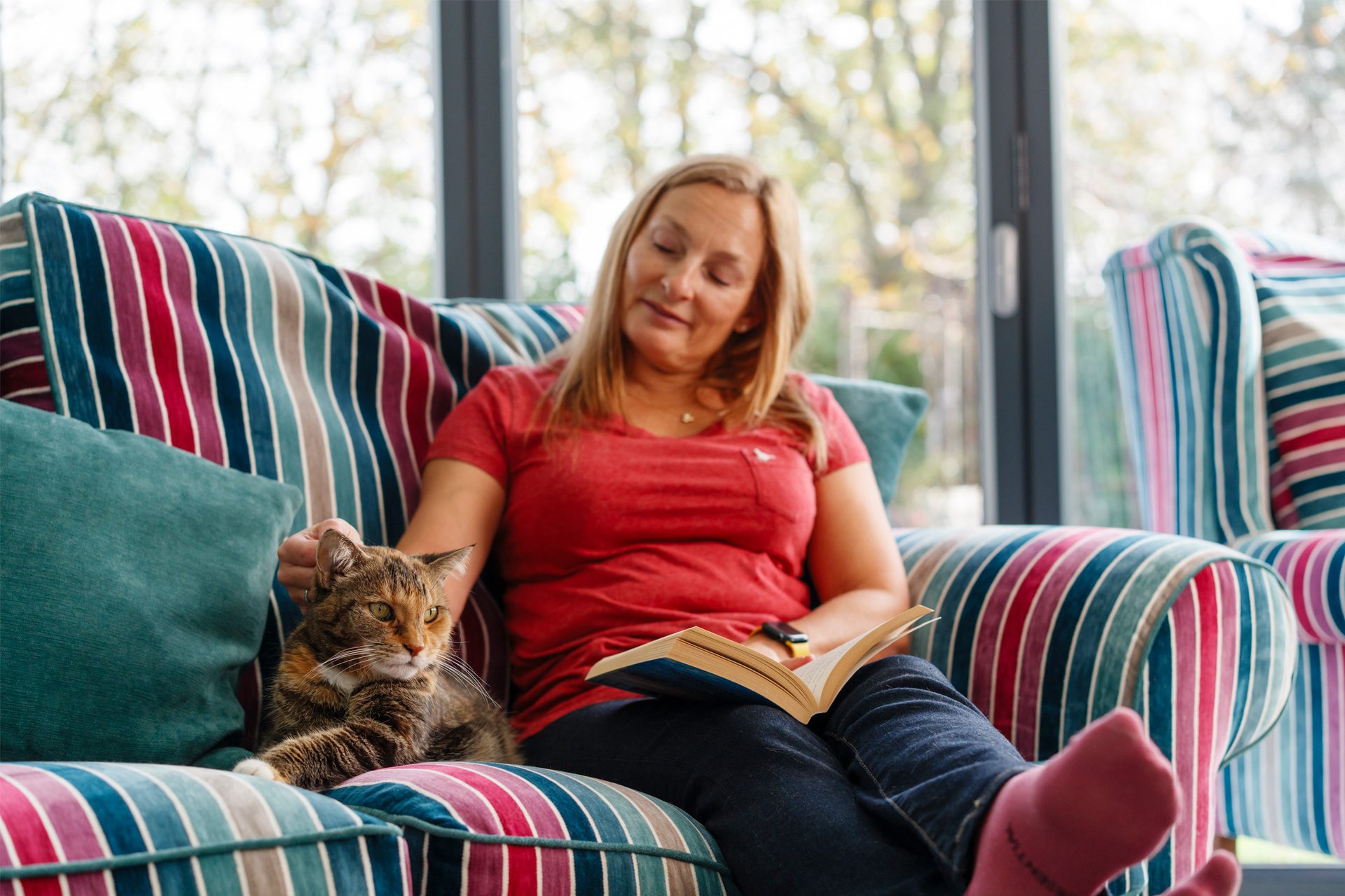 A woman relaxes on a colorful striped sofa, petting her cat while reading a book.