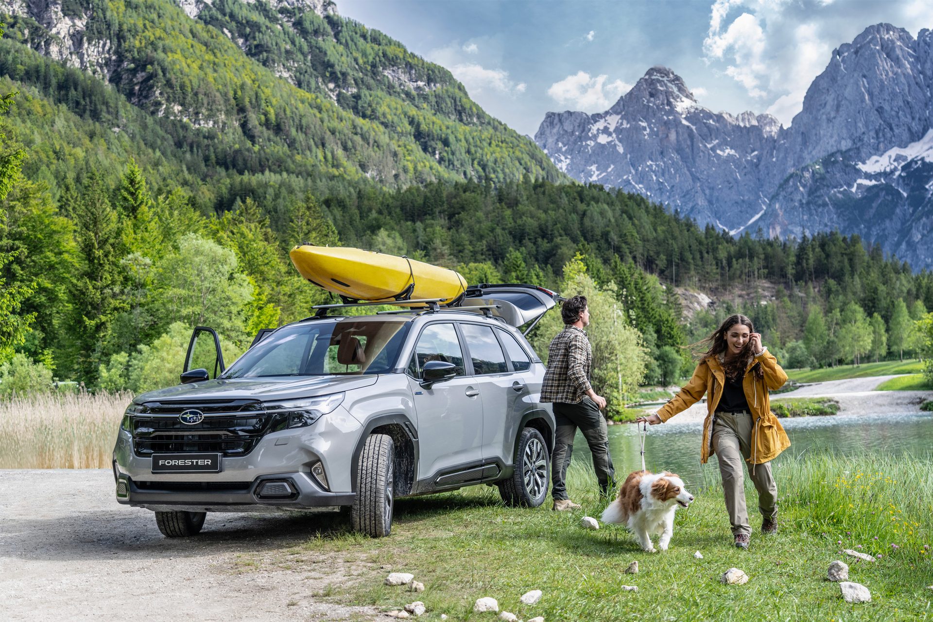 Grey Subaru Forester with kayak, couple, and dog by lake in mountains.