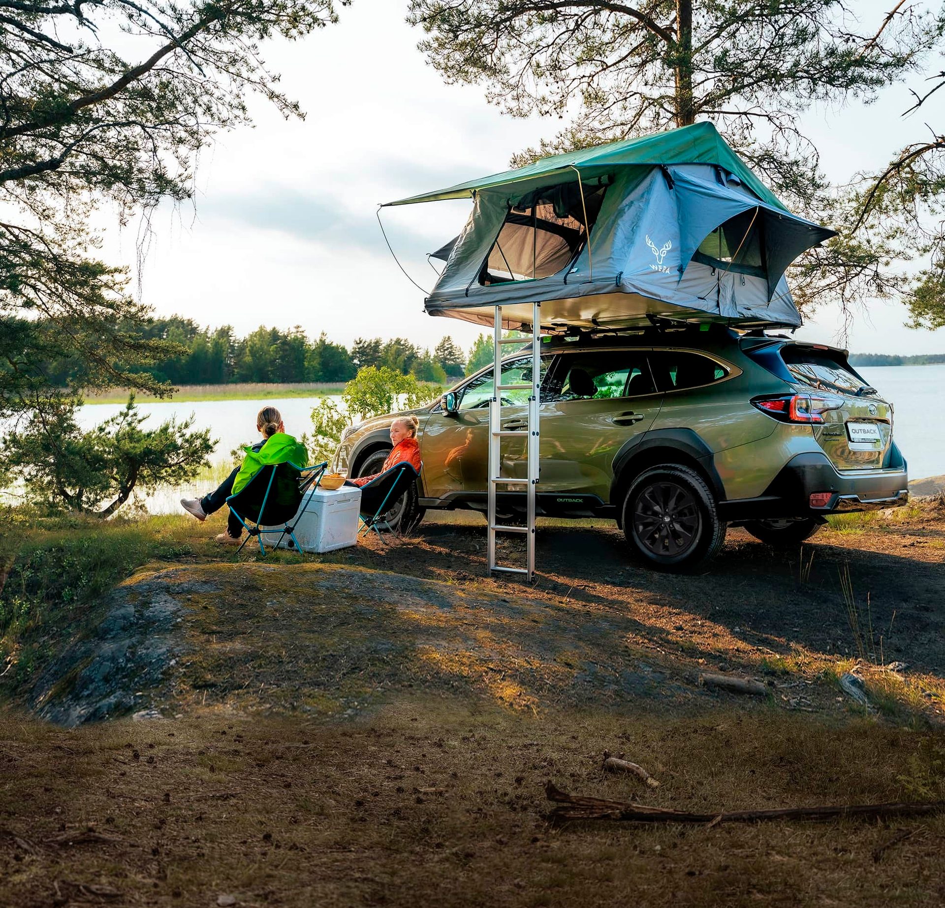 Two people camp by a lake with a car and rooftop tent.