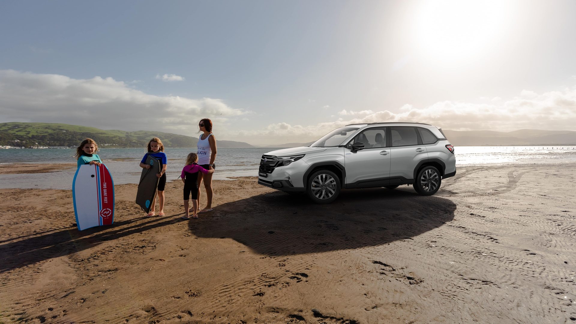 Woman, three children, and a silver SUV on a sunny beach with mountains in background.