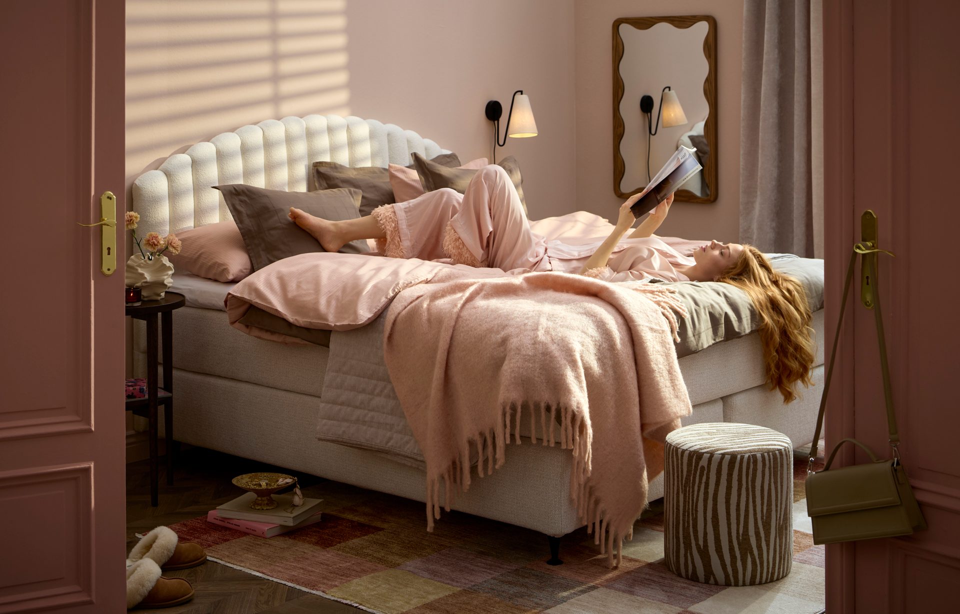 A woman reads in a sunlit pink bedroom with cozy bedding and decor.