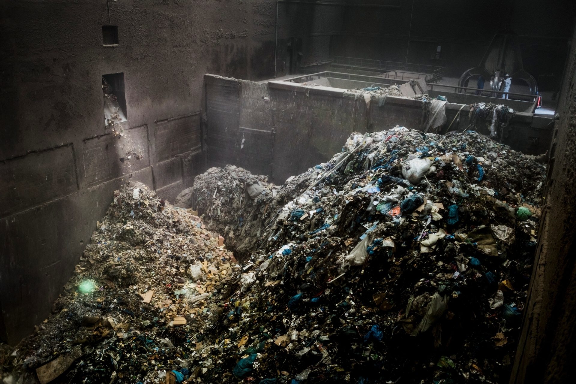 Huge piles of garbage in a dark indoor waste facility, with trash falling from a chute.