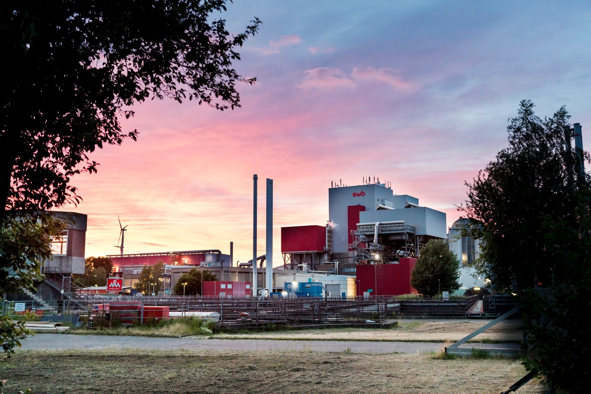 Industrial complex with "swb" and "ARA" logos under a vibrant pink/purple sunset, framed by trees, with wind turbines.