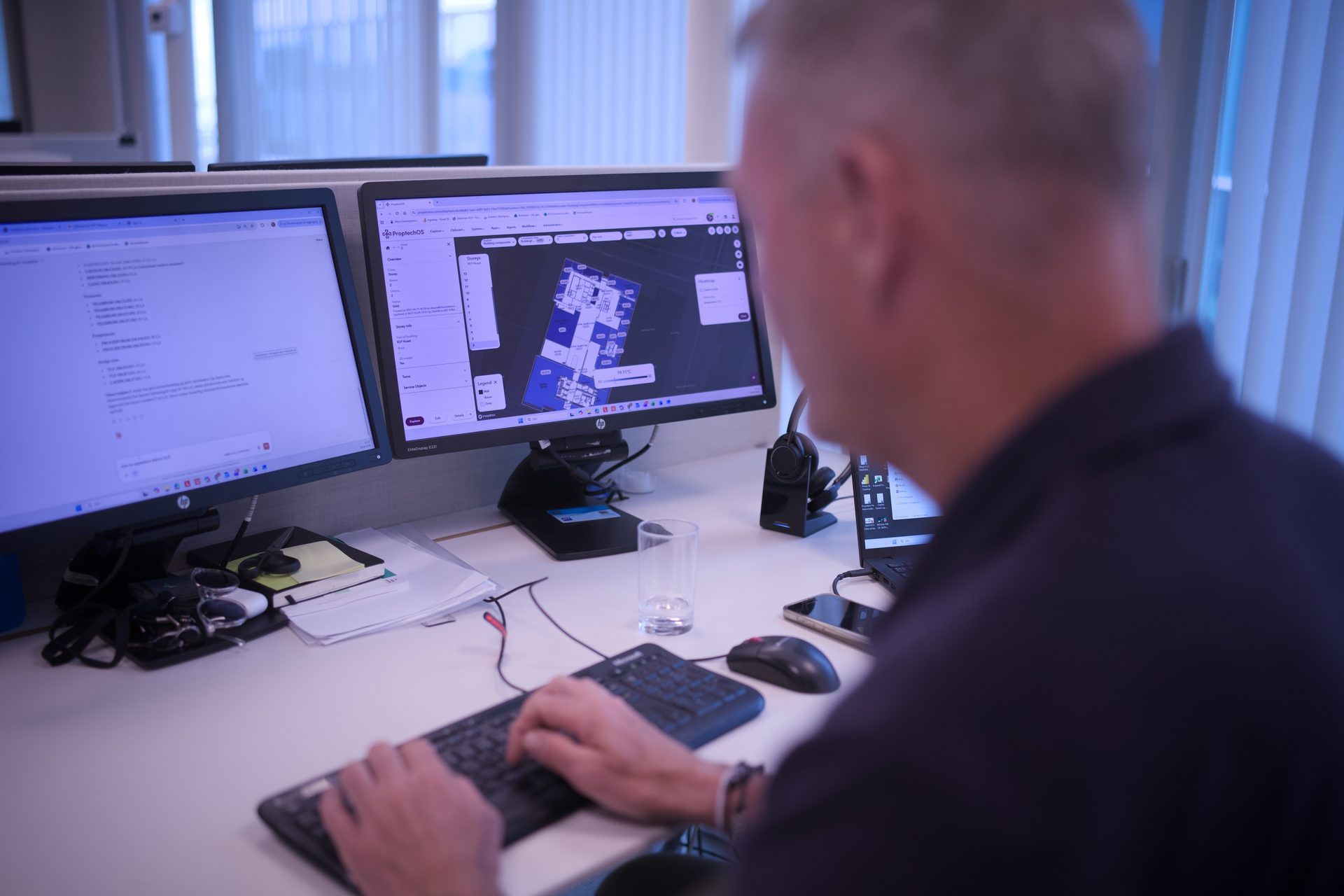 Man working at a desk with two computer monitors displaying data and a floor plan application.