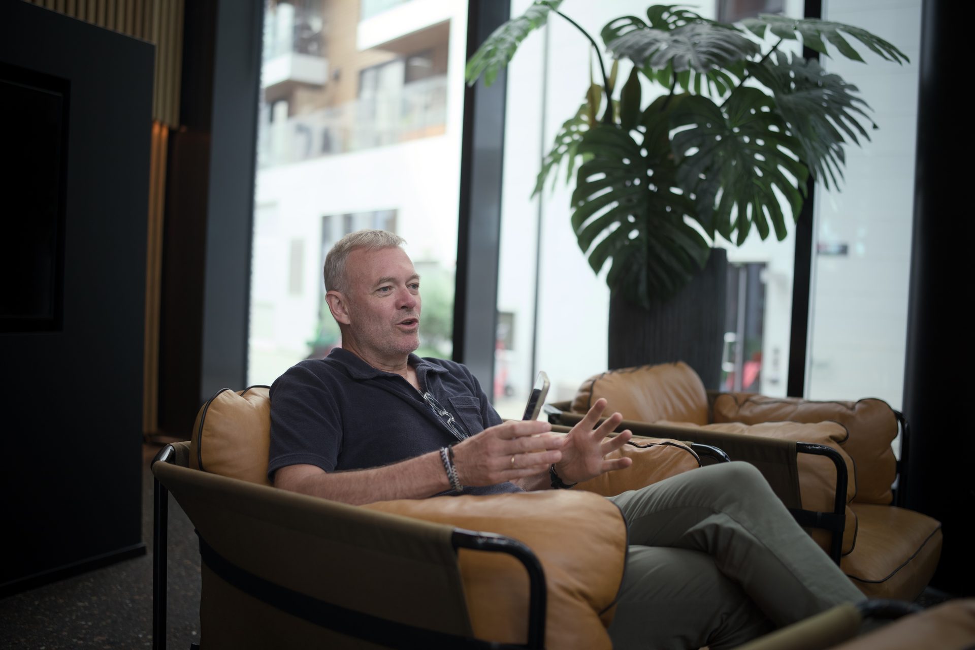 Man in a dark polo shirt gesturing while sitting in a brown leather chair, talking indoors.
