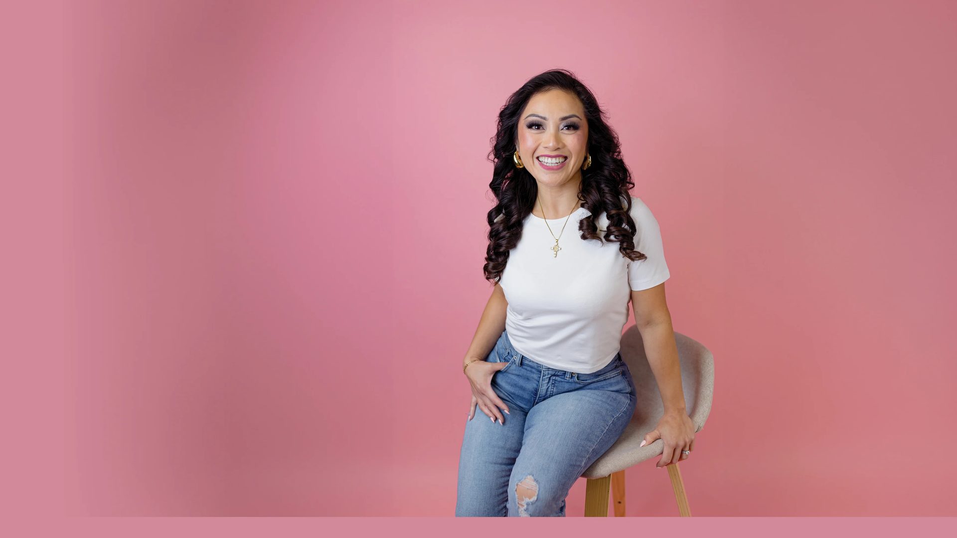 A smiling woman with dark curly hair, wearing a white t-shirt and jeans, sitting on a chair against a pink background.
