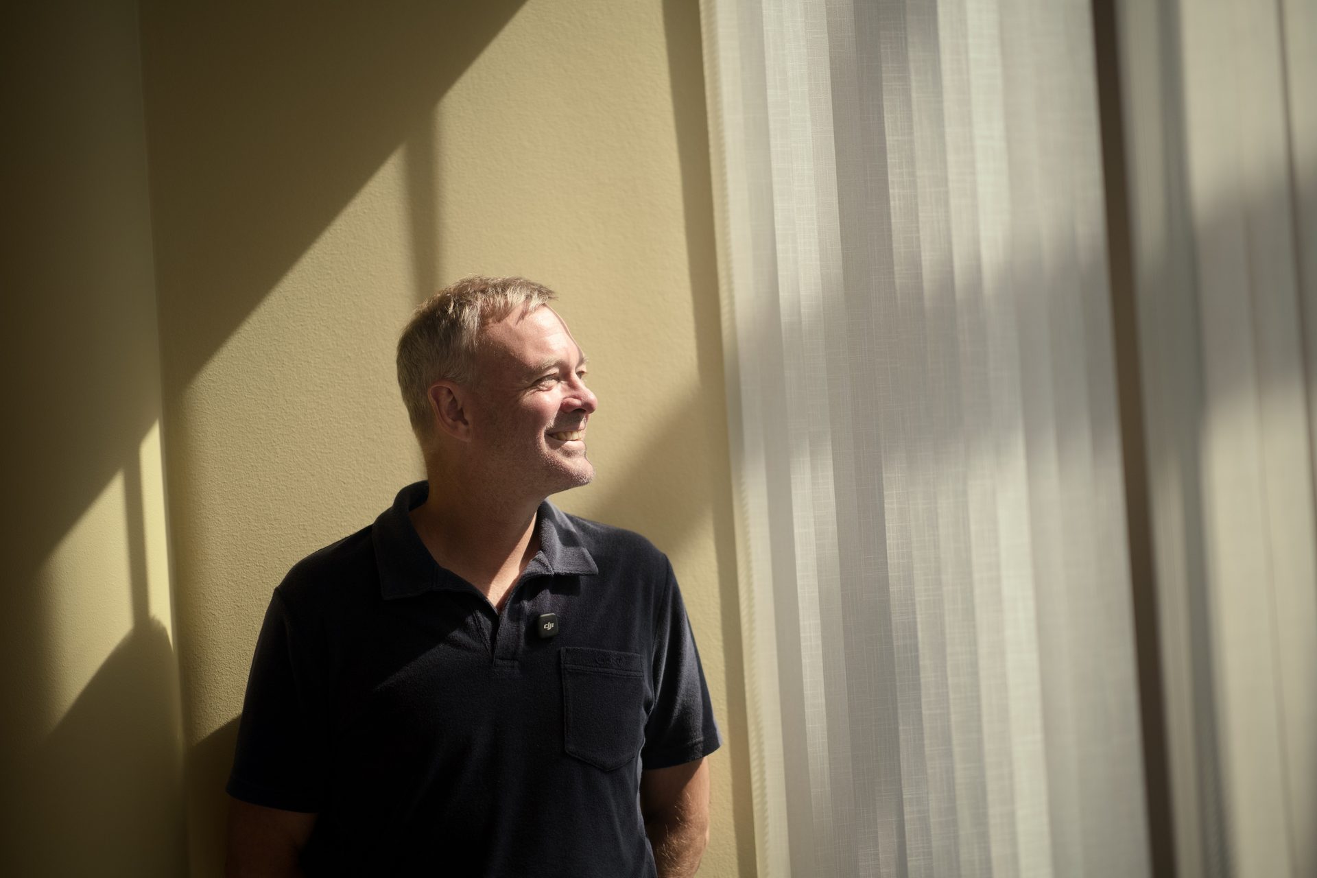 A smiling man with gray hair in a dark polo shirt looks towards light from a window.