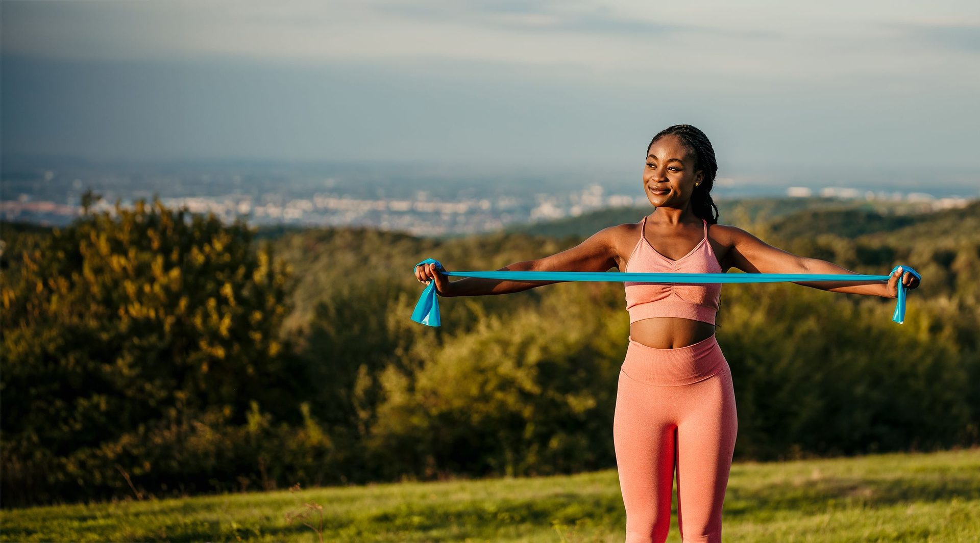 A woman in athletic wear exercising outdoors with a blue resistance band, smiling, with a scenic view.