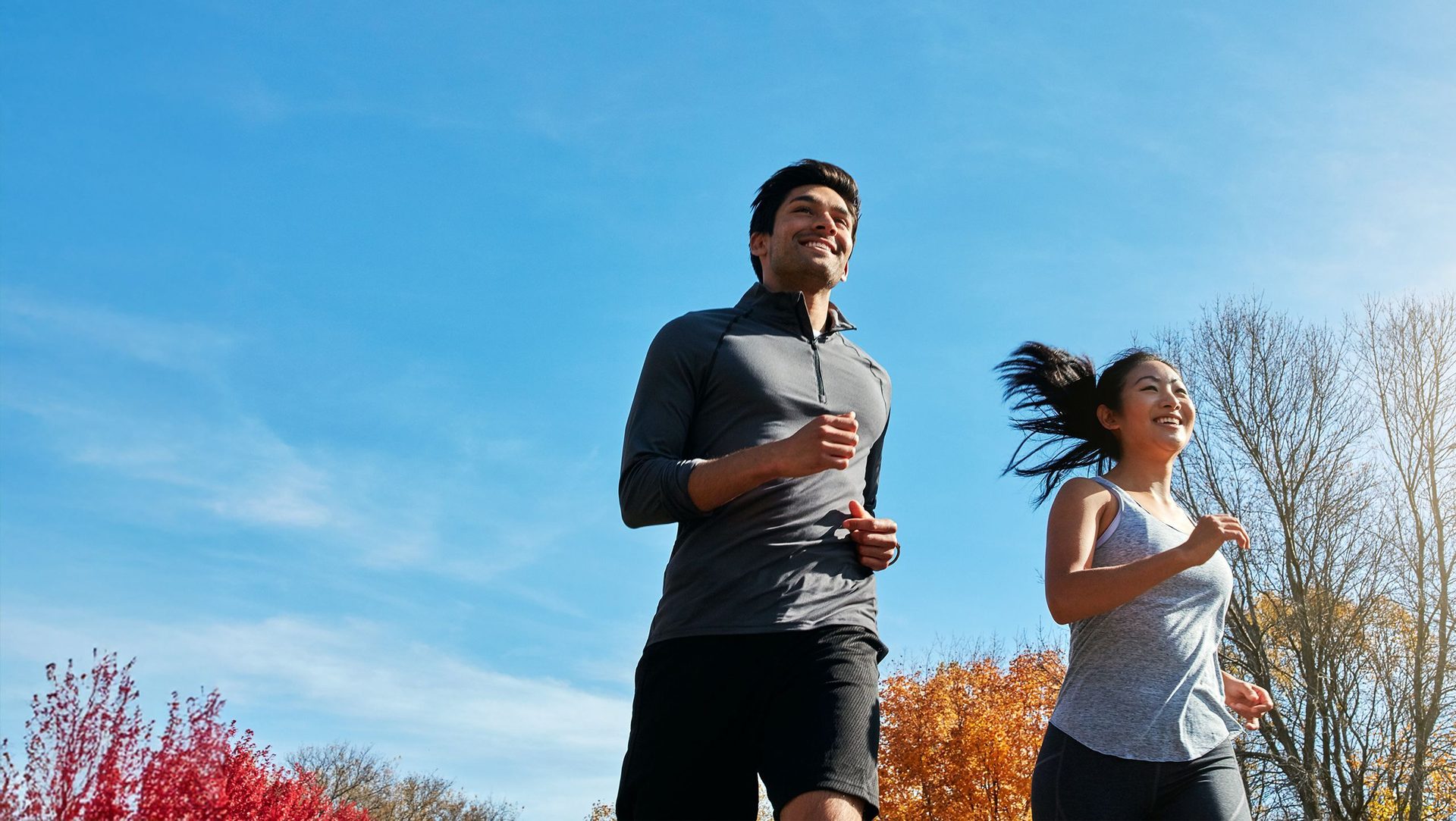 Two smiling people, a man and a woman, jogging outdoors under a clear blue sky in an autumn park.