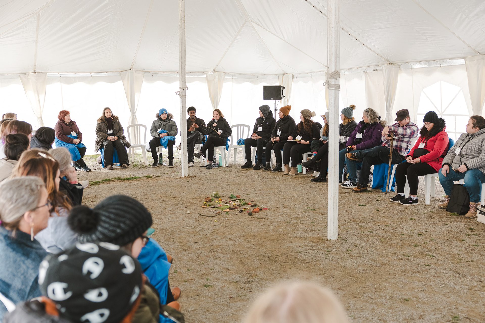 A circle of early childhood educators seated in a tent, having a discussion. In the centre of the circle, there is an assortment of rocks, leaves and sticks.  