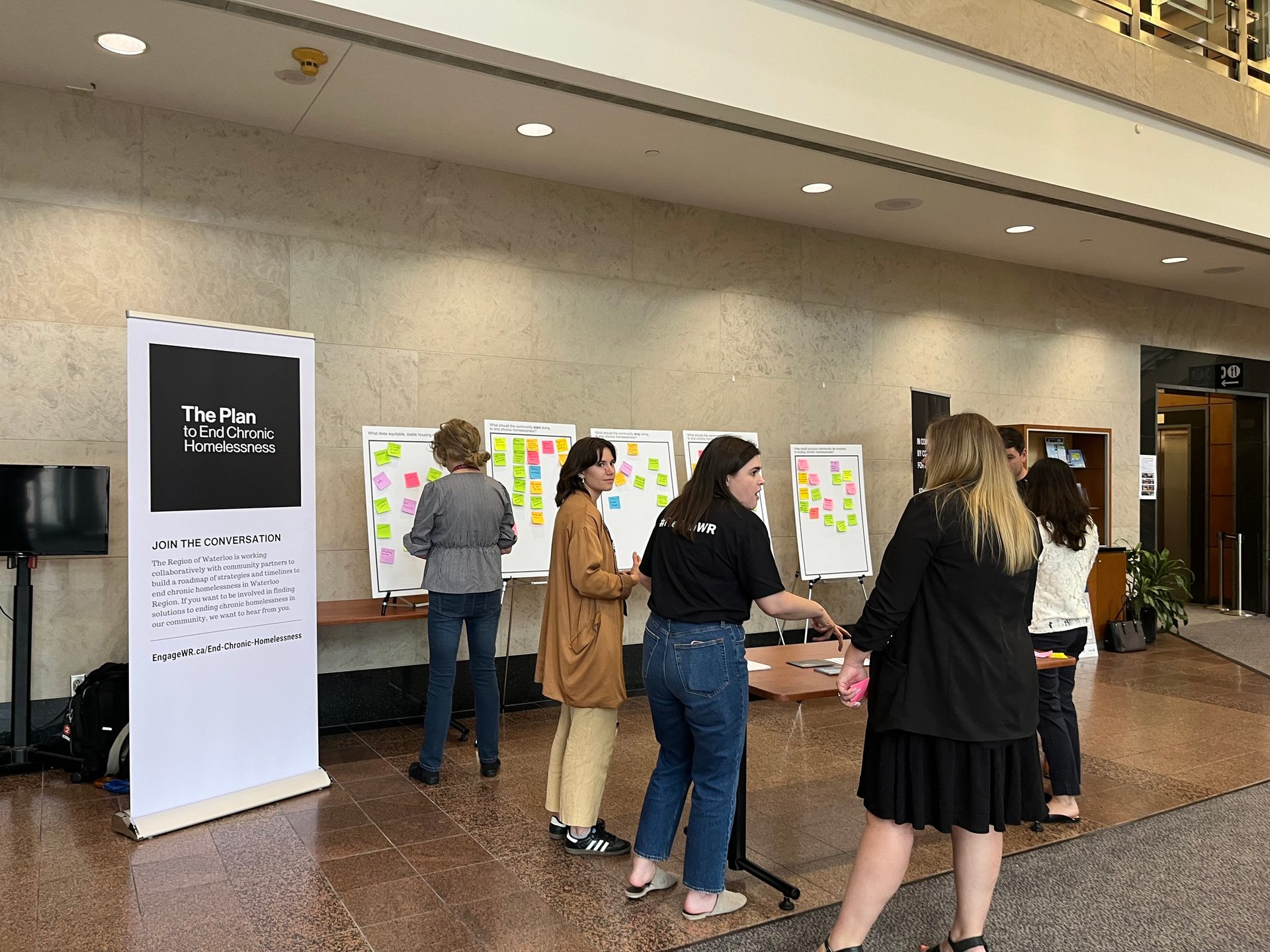Several people are interacting around an information booth in the lobby of a Region of Waterloo building. The information booth has a stand-up banner with a logo for the Plan to End Chronic Homelessness. One person is placing sticky notes on an interactive board to collect feedback. 