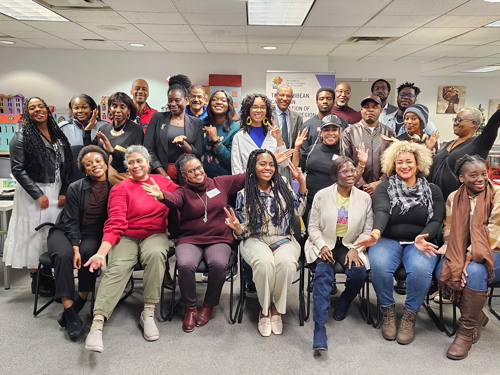 A large group of smiling Black adults, some standing and some seated, wave at the camera indoors.