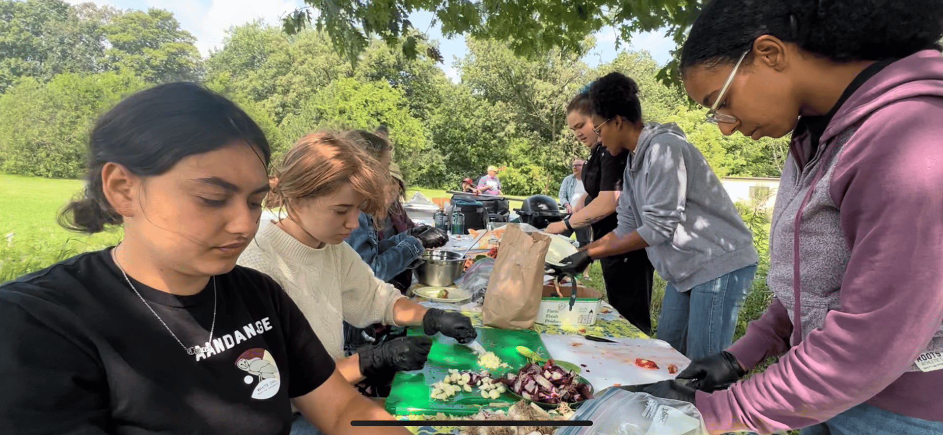 A group of young women outdoors, wearing gloves, chopping vegetables on a table.
