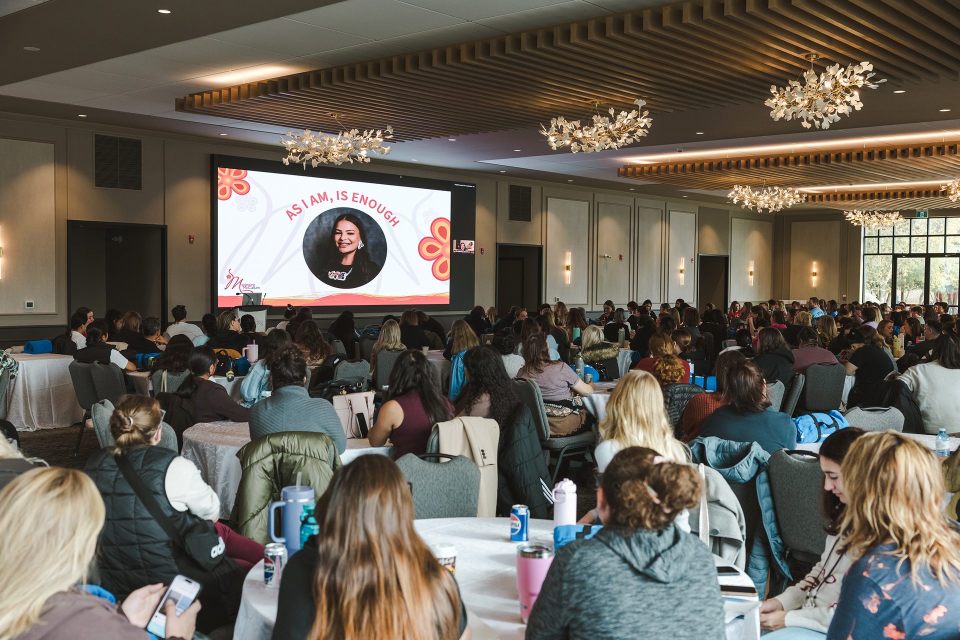 A room full of early childhood educators who are looking at a slide show in a conference room. The slide show features a photo of the Indigenous speaker and the words “As I am, Is Enough” 