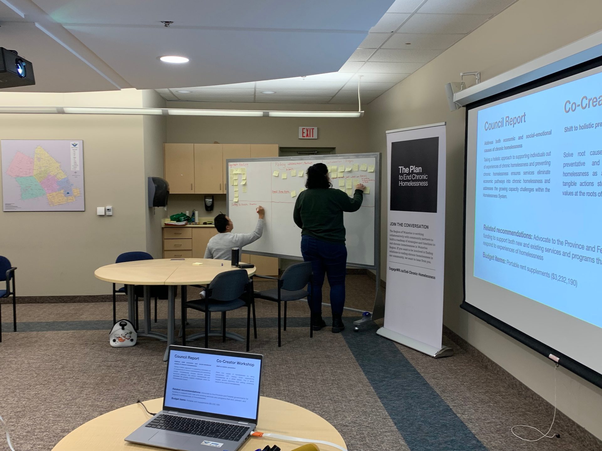 Two people place sticky notes on a whiteboard at a PECH Co-creators meeting. They are in a meeting room.