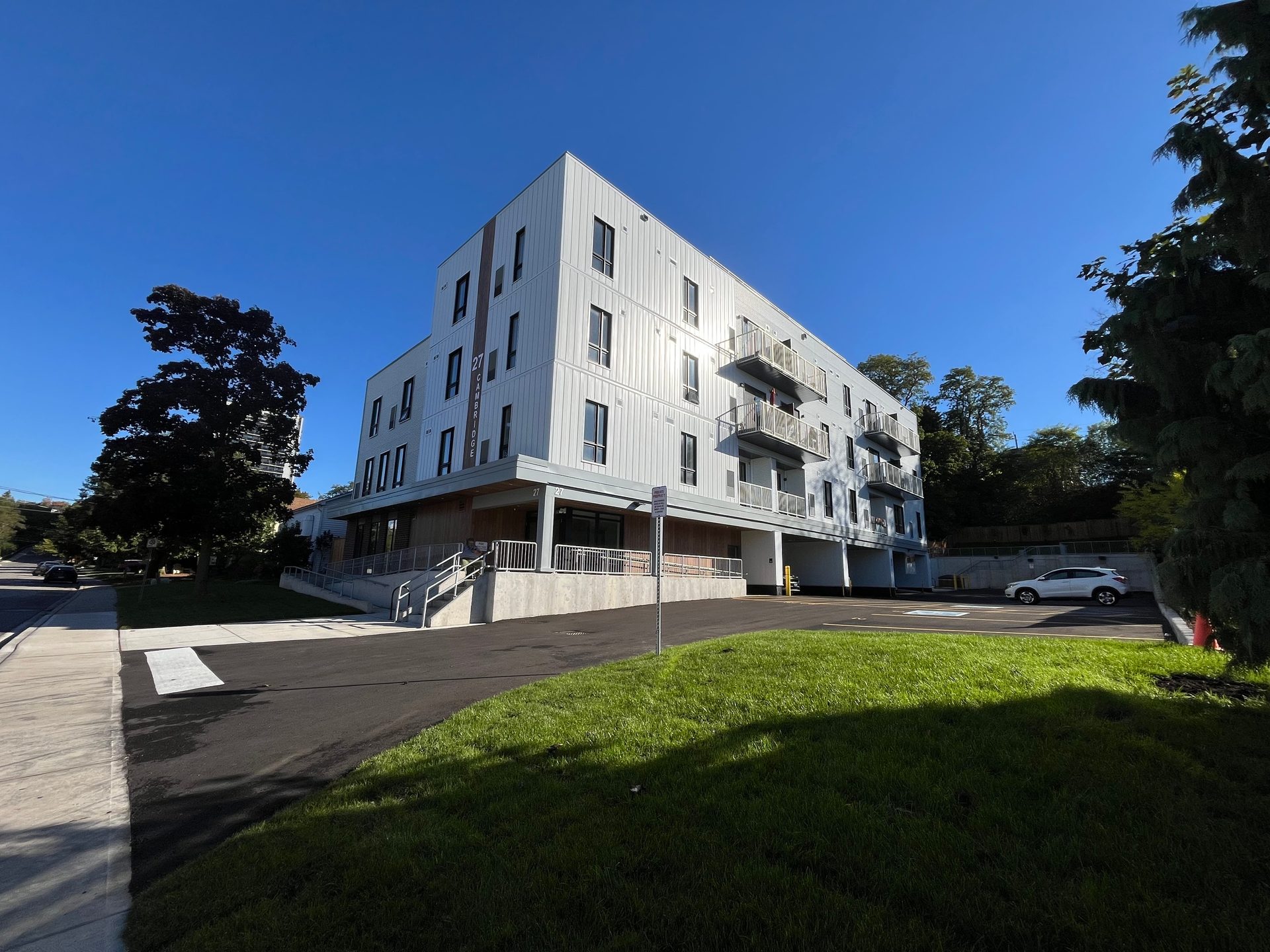 An exterior view of a transitional housing development on a residential street in Cambridge. It's a modern-looking multi-residential building with four floors, balconies, a parking lot, and green grass under a clear blue sky.