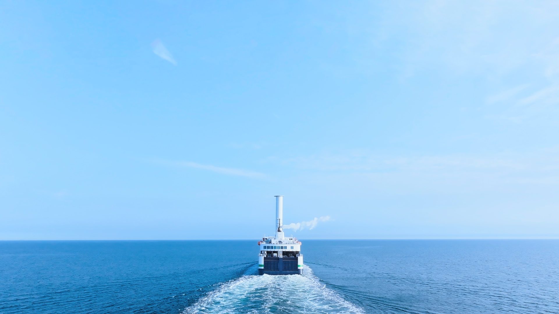 A large ferry boat sails away across a calm blue ocean, leaving a white wake under a clear sky.