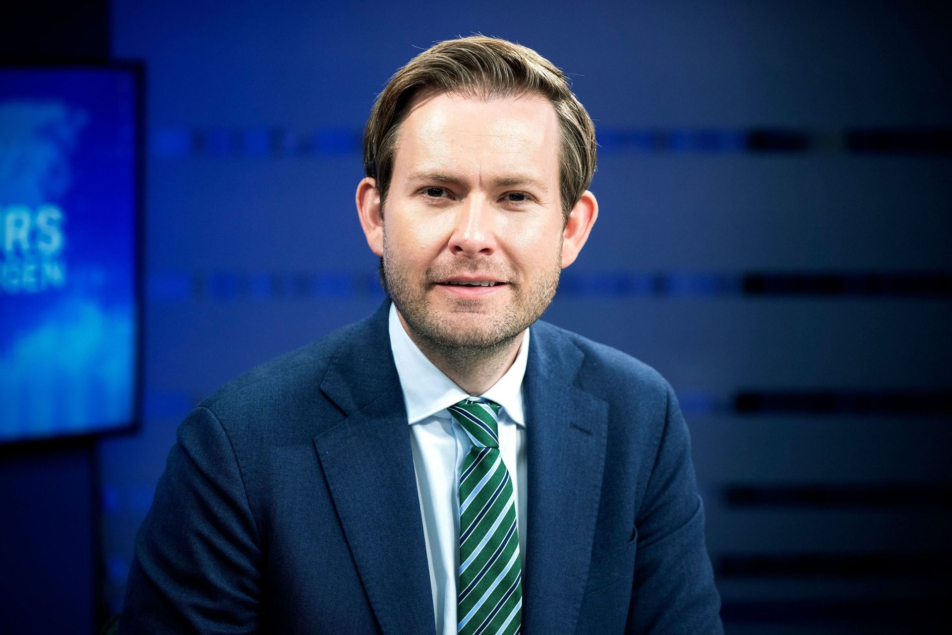 A man in a blue suit and striped tie, smiling confidently in front of a blurred blue background.