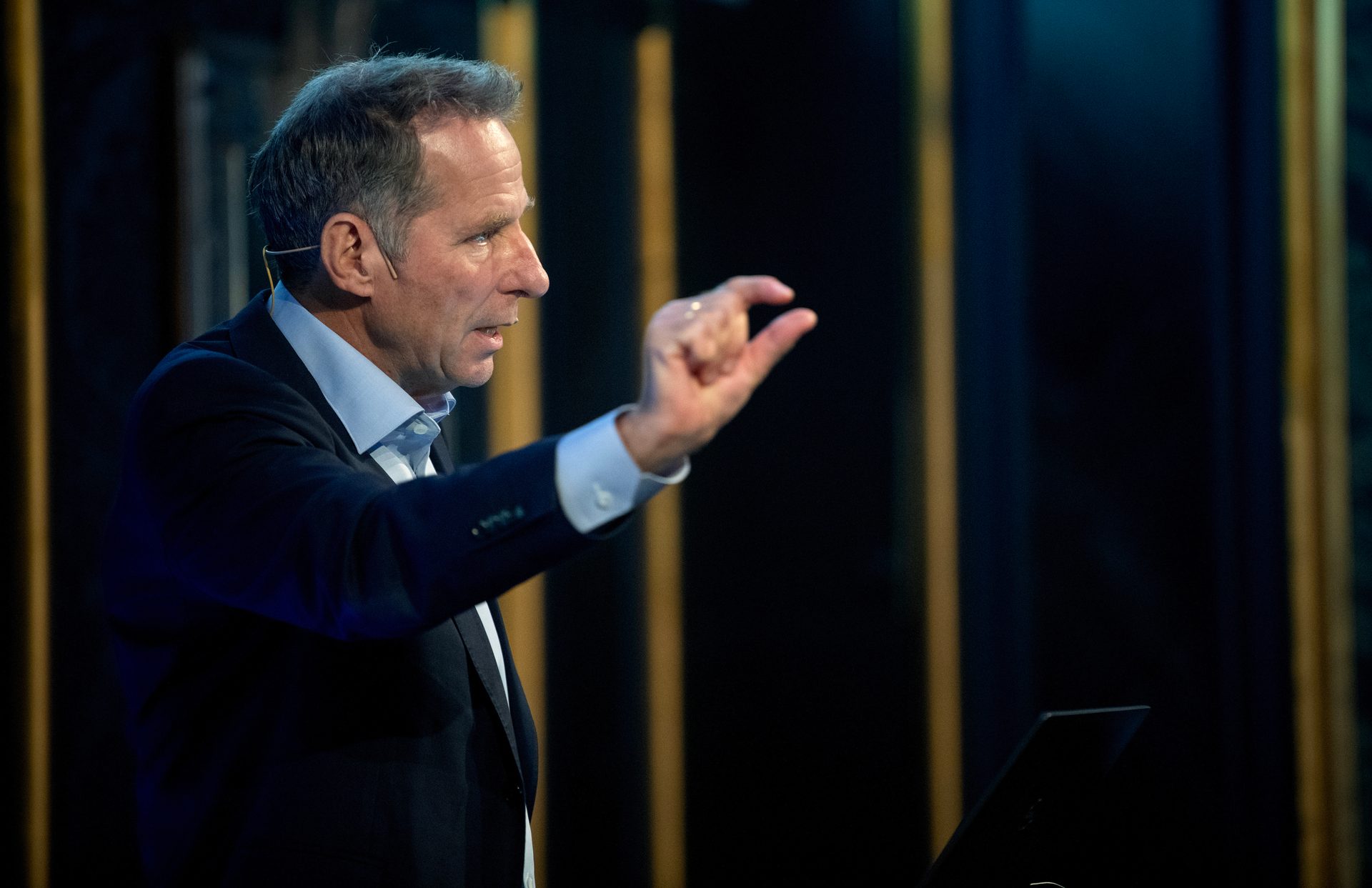Man in a suit gesturing with his hand, speaking on stage with dark background.