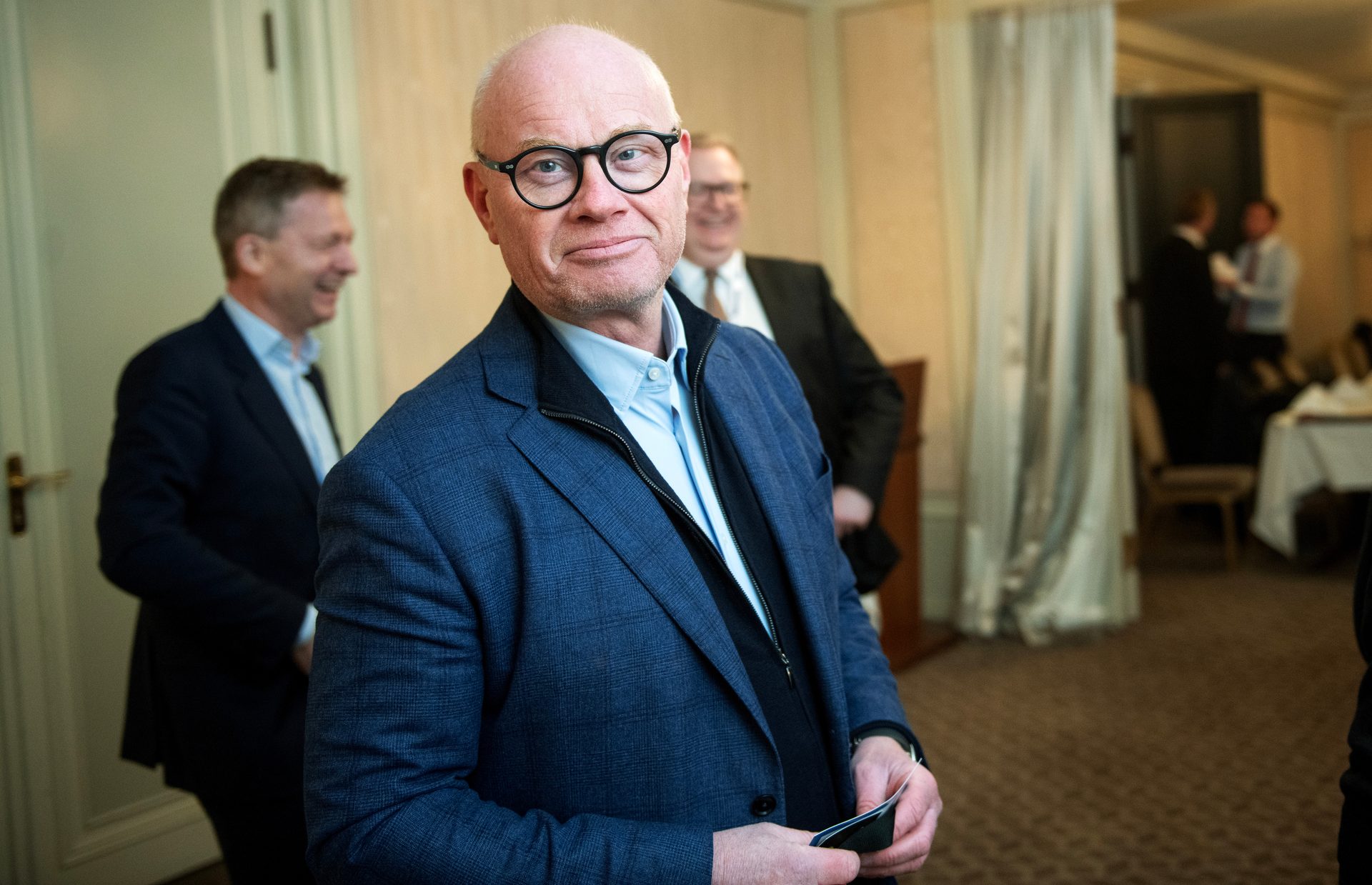 A bald man in a blue jacket and glasses smiles slightly, holding items, with blurred figures in the background.