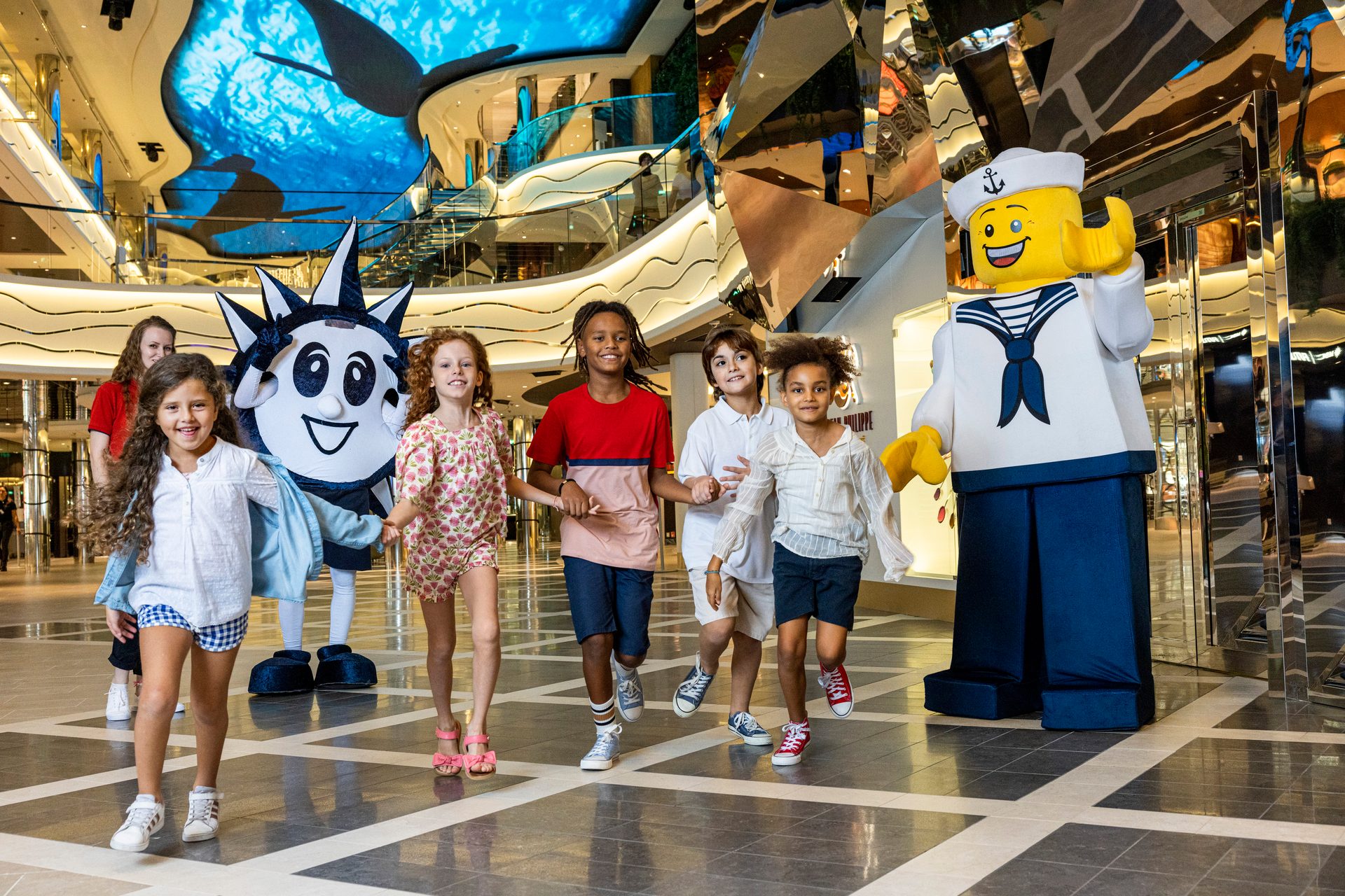 Kids play with a Lego sailor and a spiky mascot in a cruise ship atrium.