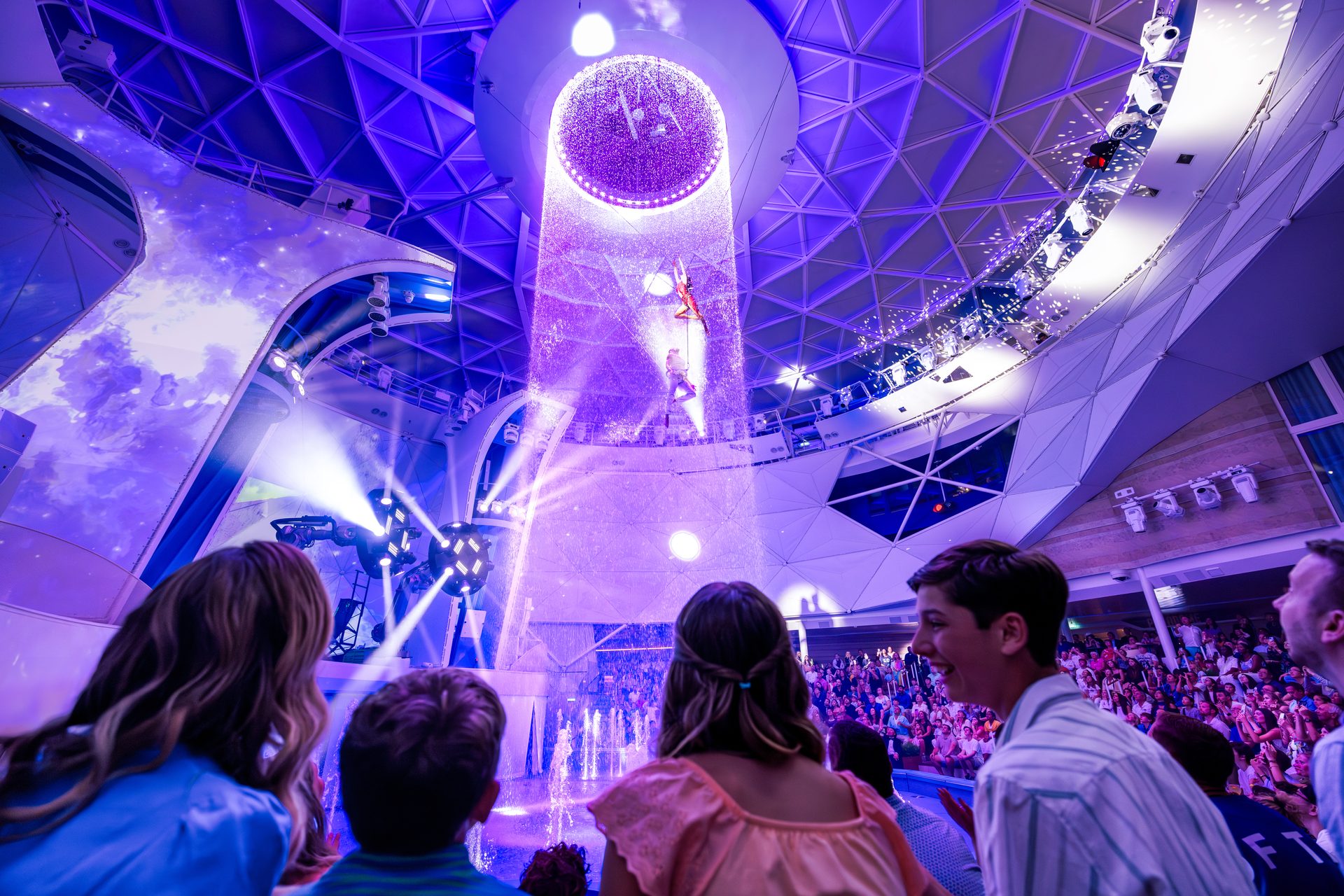 Acrobats perform in a dazzling purple water show watched by an audience.