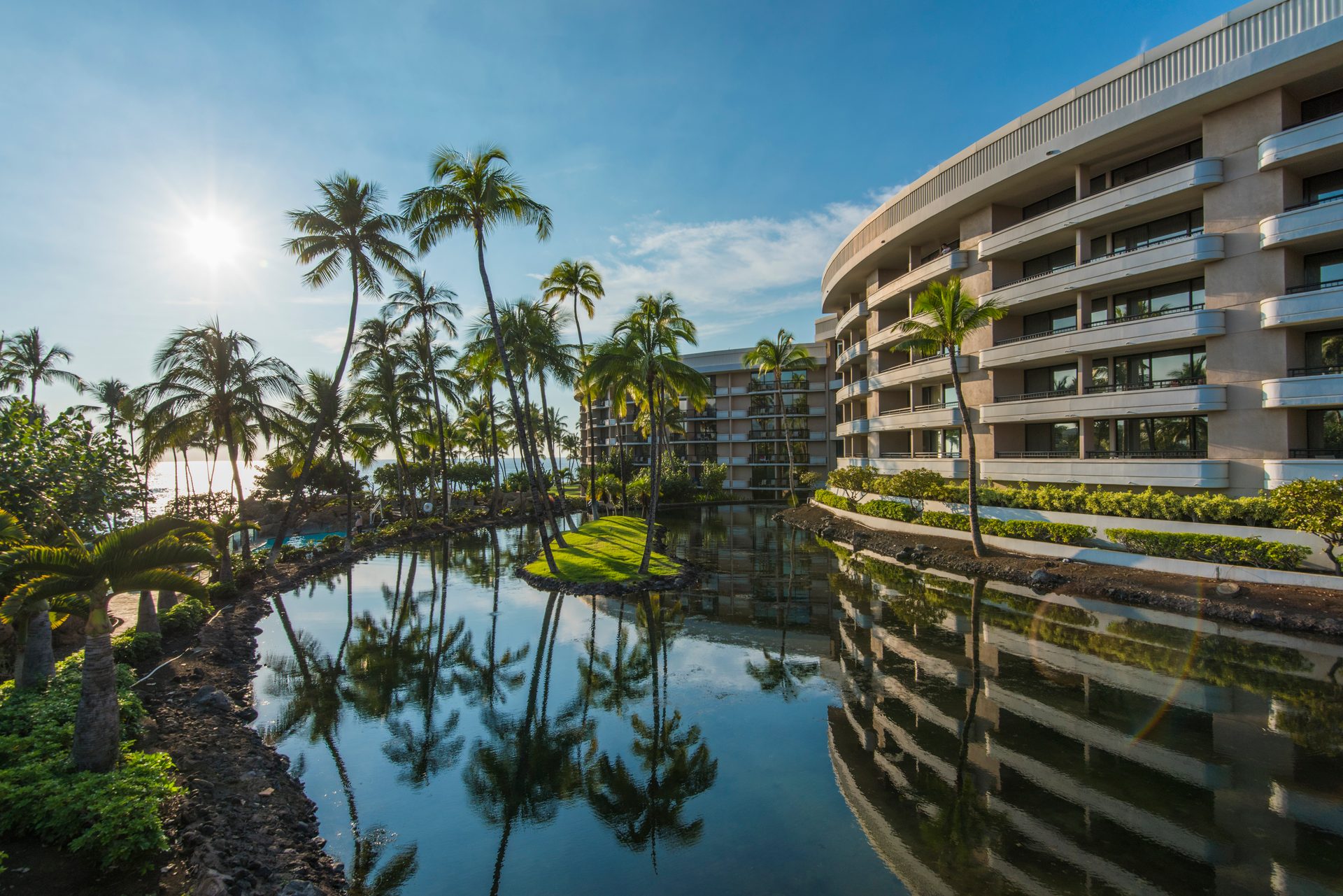 A luxurious resort hotel with a curved building, surrounded by palm trees and a reflective lagoon under a bright sun.