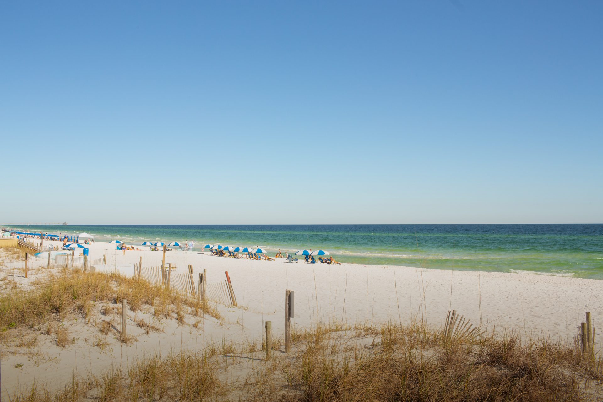Sunny white sand beach with turquoise ocean, blue sky, beach umbrellas, and dunes.