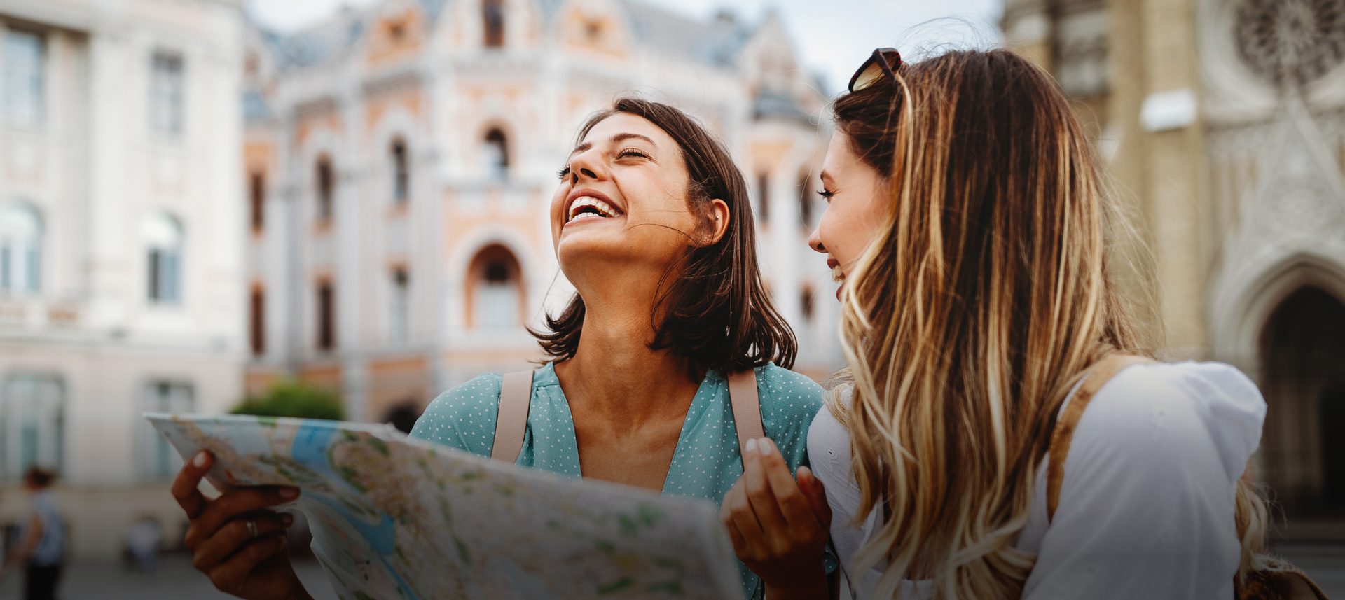 Two happy young women travelers looking at a map and laughing in a city square.