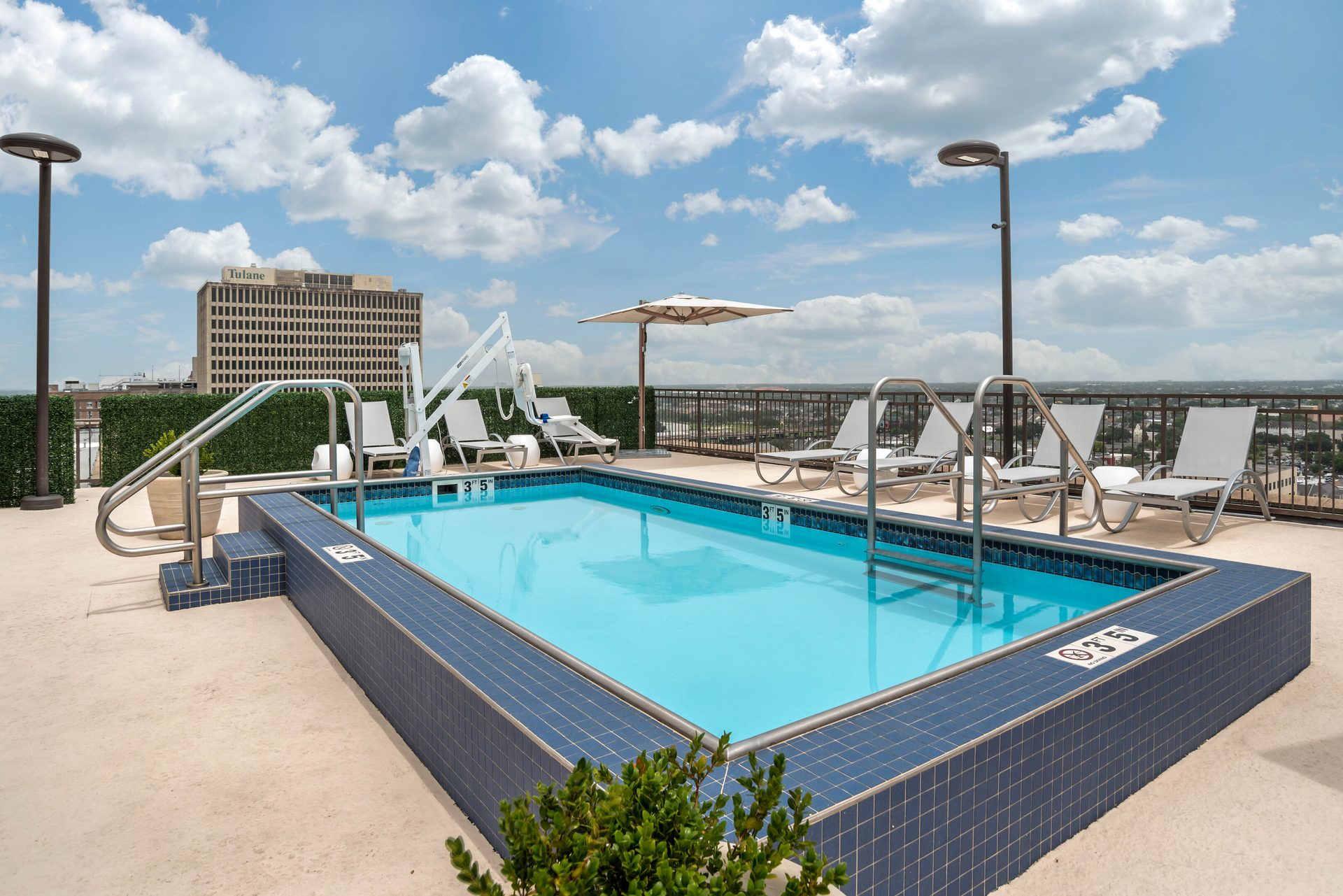 Rooftop pool with lounge chairs, a lift, and distant city buildings under a partly cloudy sky.