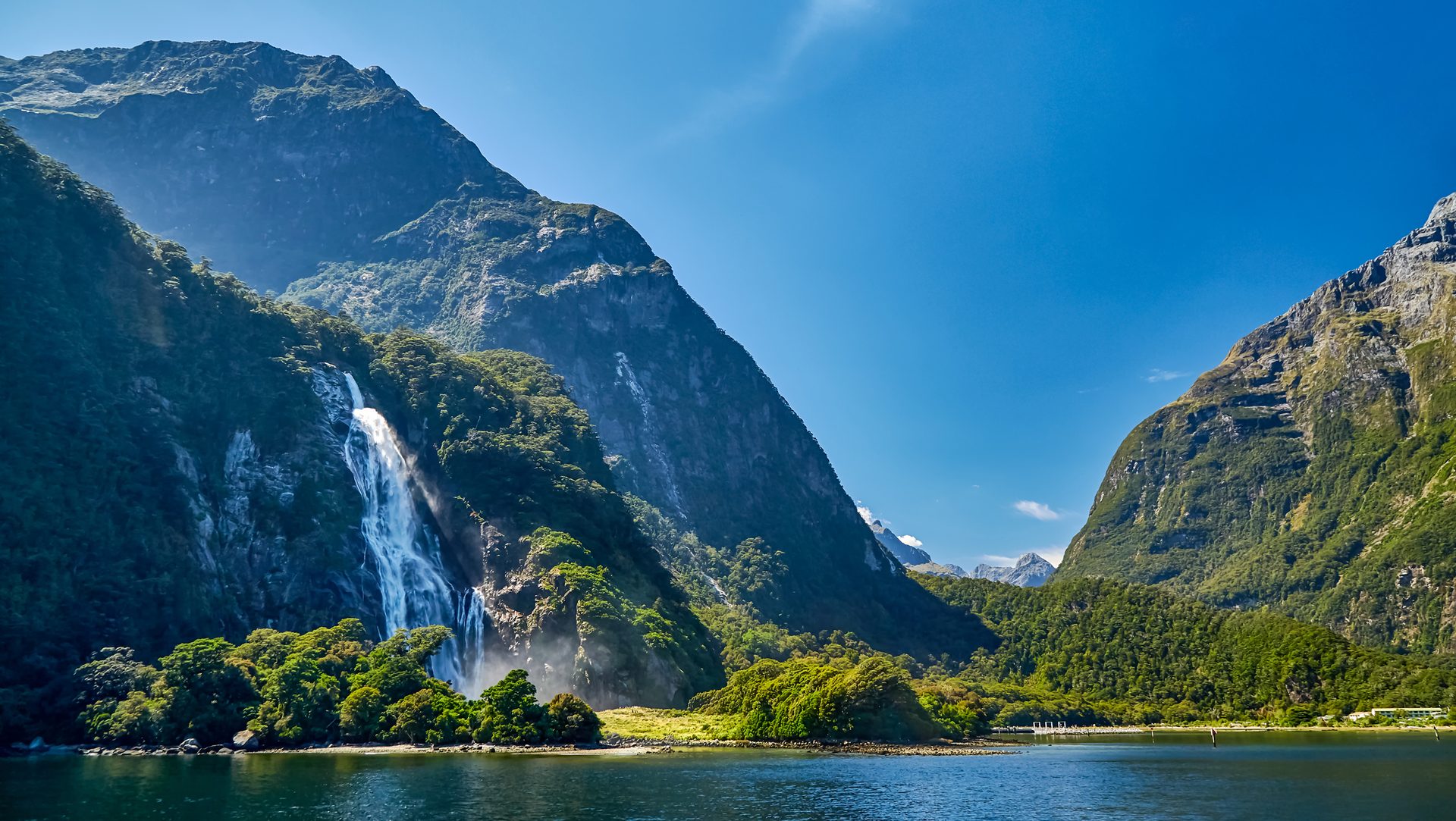 Waterfall cascades down a green mountain into a fjord under a clear sky.