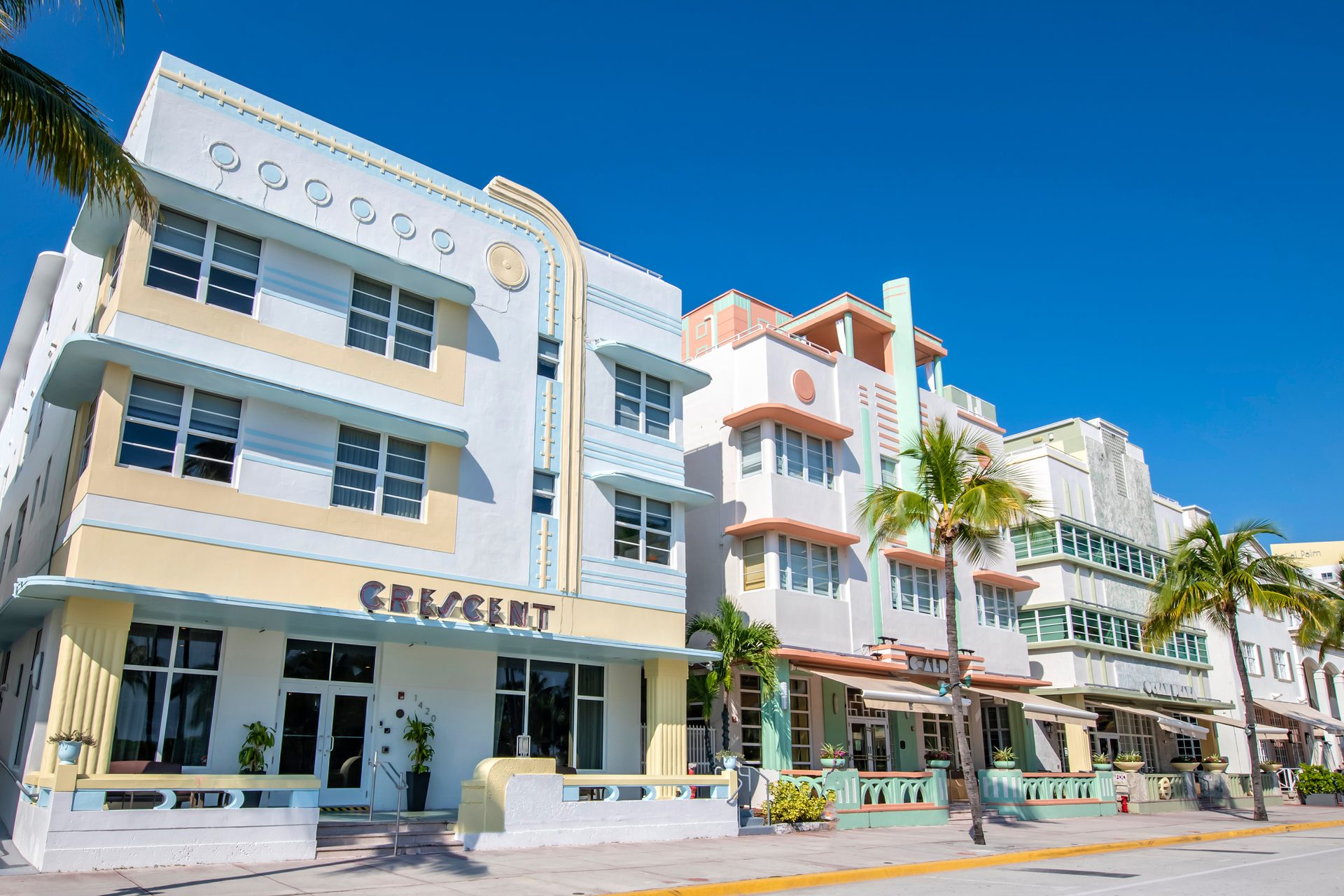 Pastel Art Deco hotels line a sunny Miami Beach street with geometric details and palm trees.