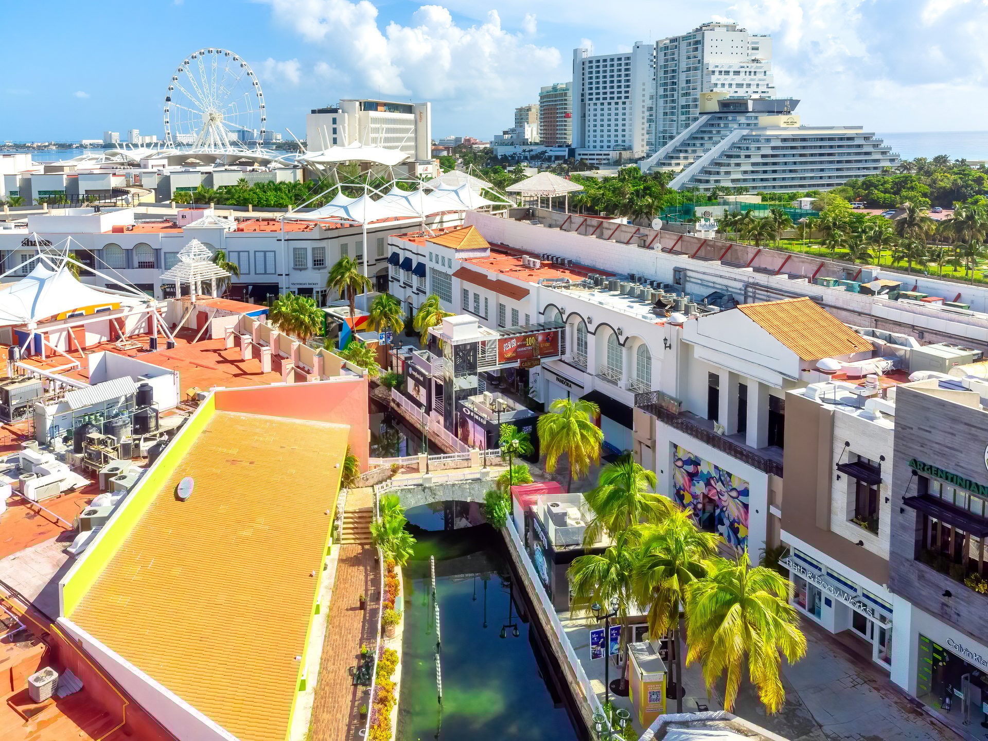 Vibrant aerial view of a shopping complex with a canal, palm trees, a Ferris wheel, and hotels.