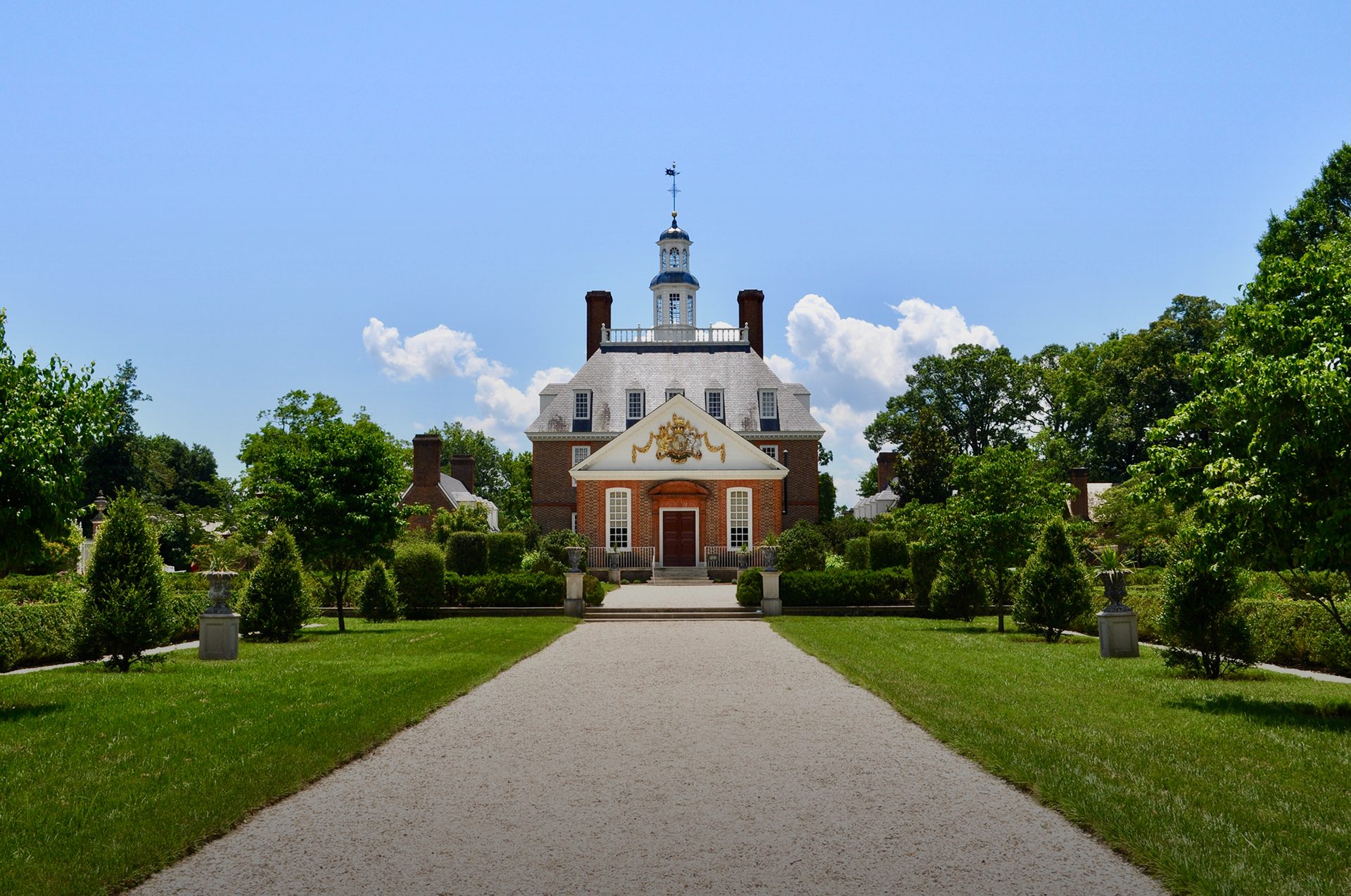 Grand brick mansion with a cupola, centered on a gravel path through manicured gardens, blue sky.