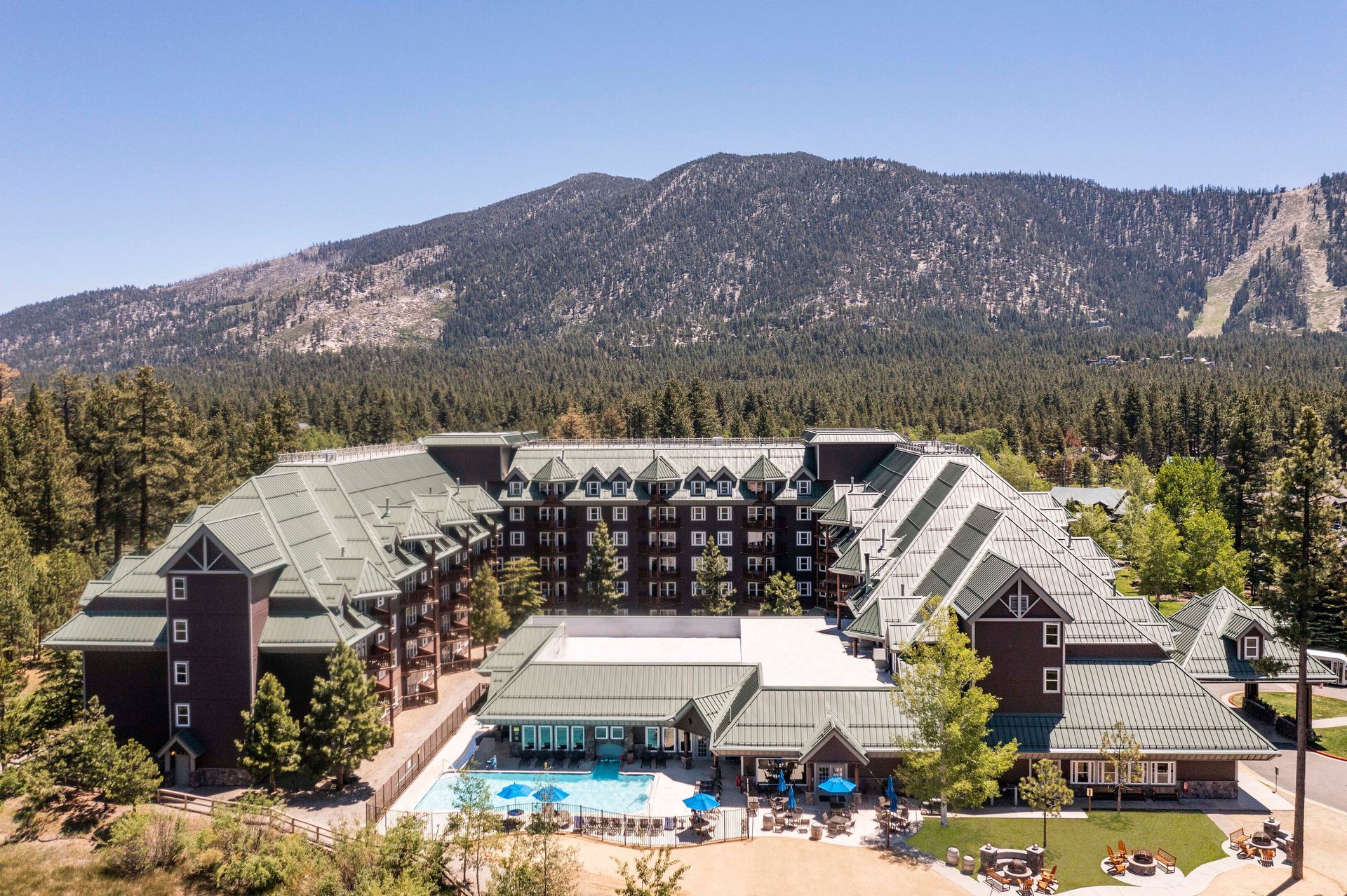 Aerial view of a large resort with green roofs and outdoor pool, surrounded by pine forests and mountains.