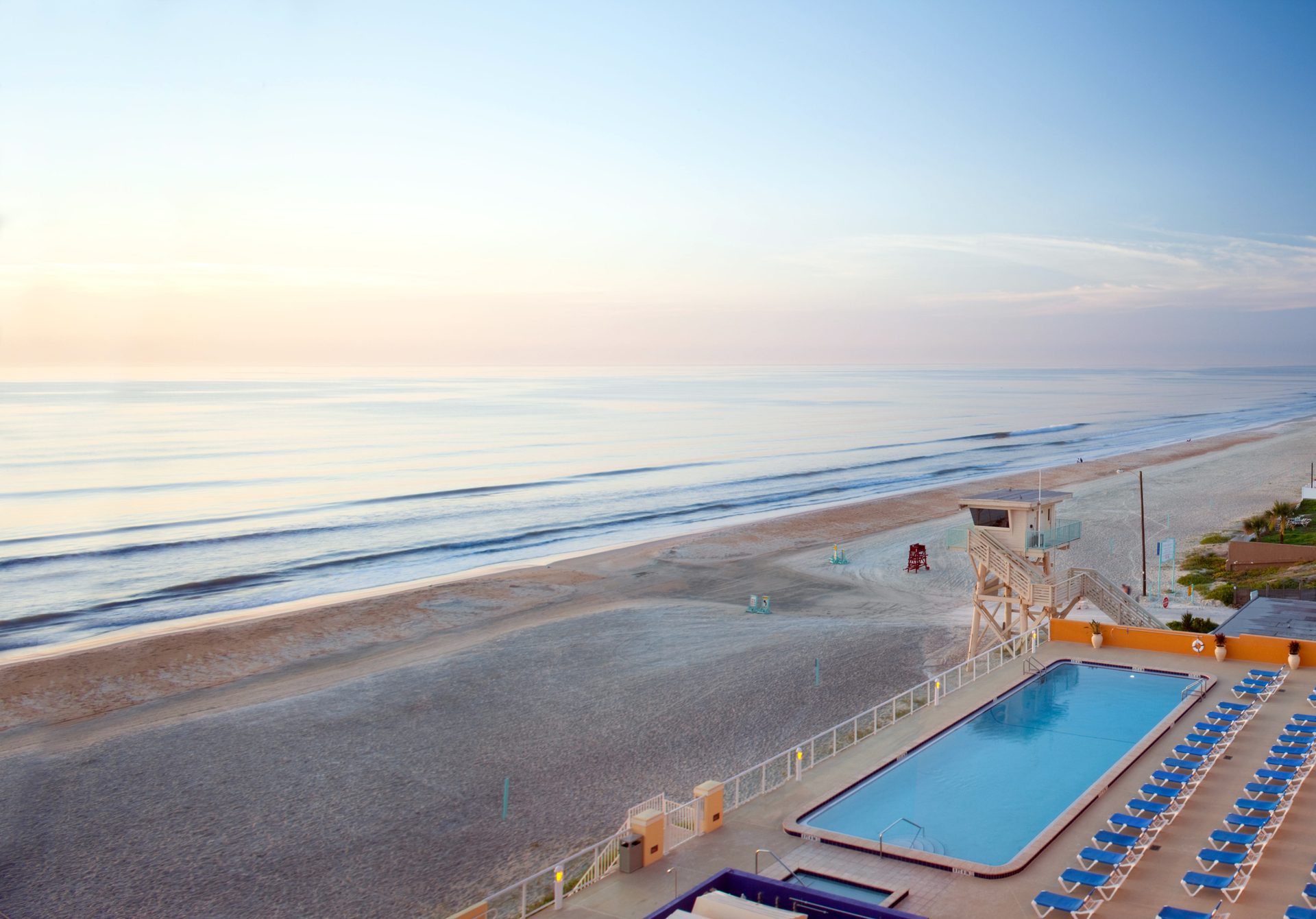 Aerial view of a sandy beach, ocean waves, a lifeguard tower, and a resort swimming pool.