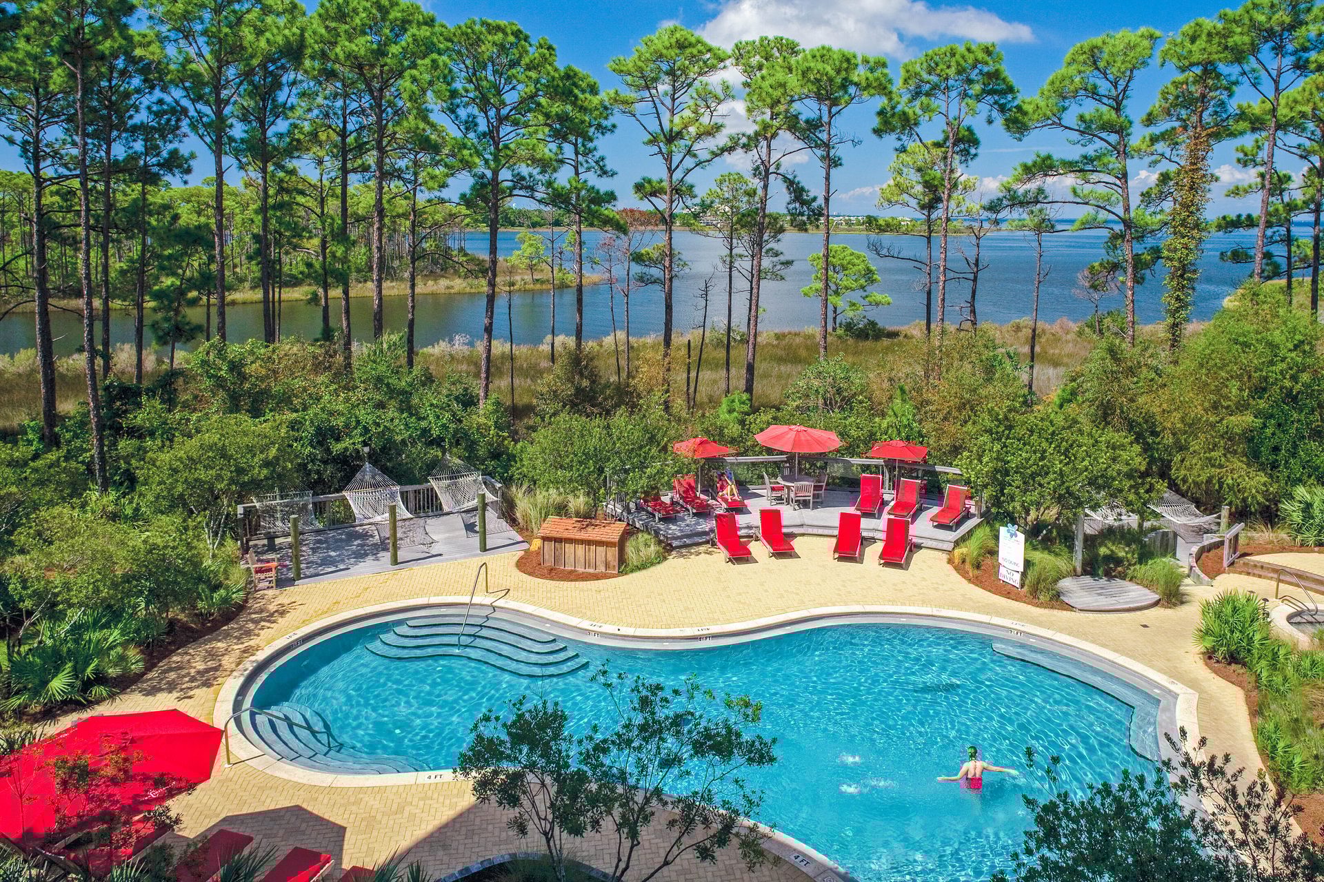Resort pool with red lounge chairs, hammocks, and a lake surrounded by tall pine trees.