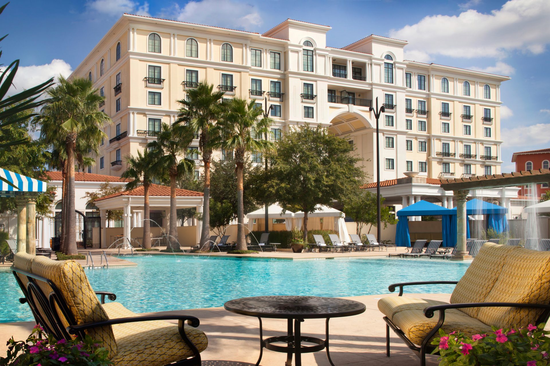 Resort hotel overlooking a large swimming pool with palm trees and lounge seating on a sunny day.