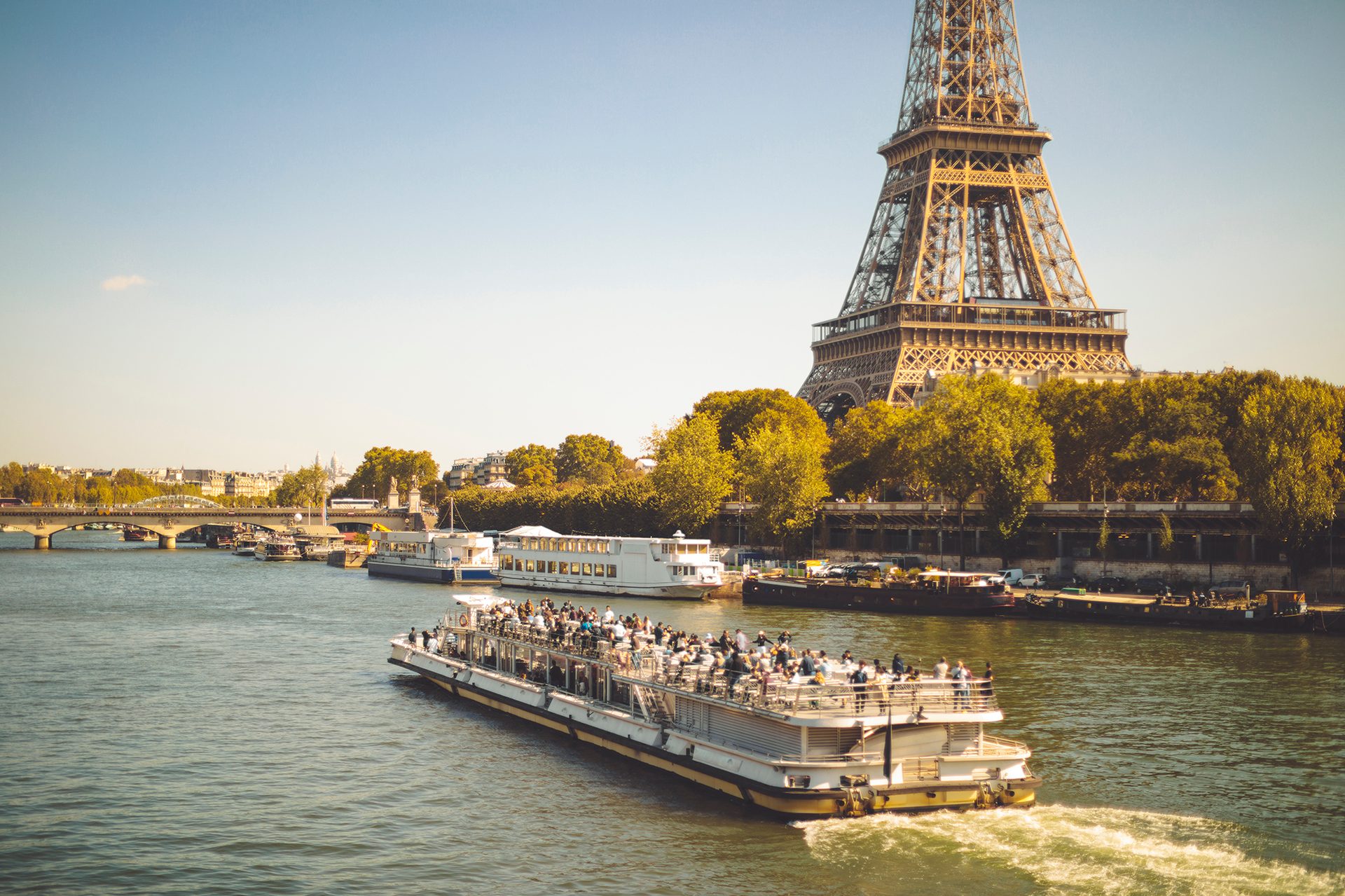 Eiffel Tower overlooks the Seine River with a tour boat full of people and Parisian buildings.